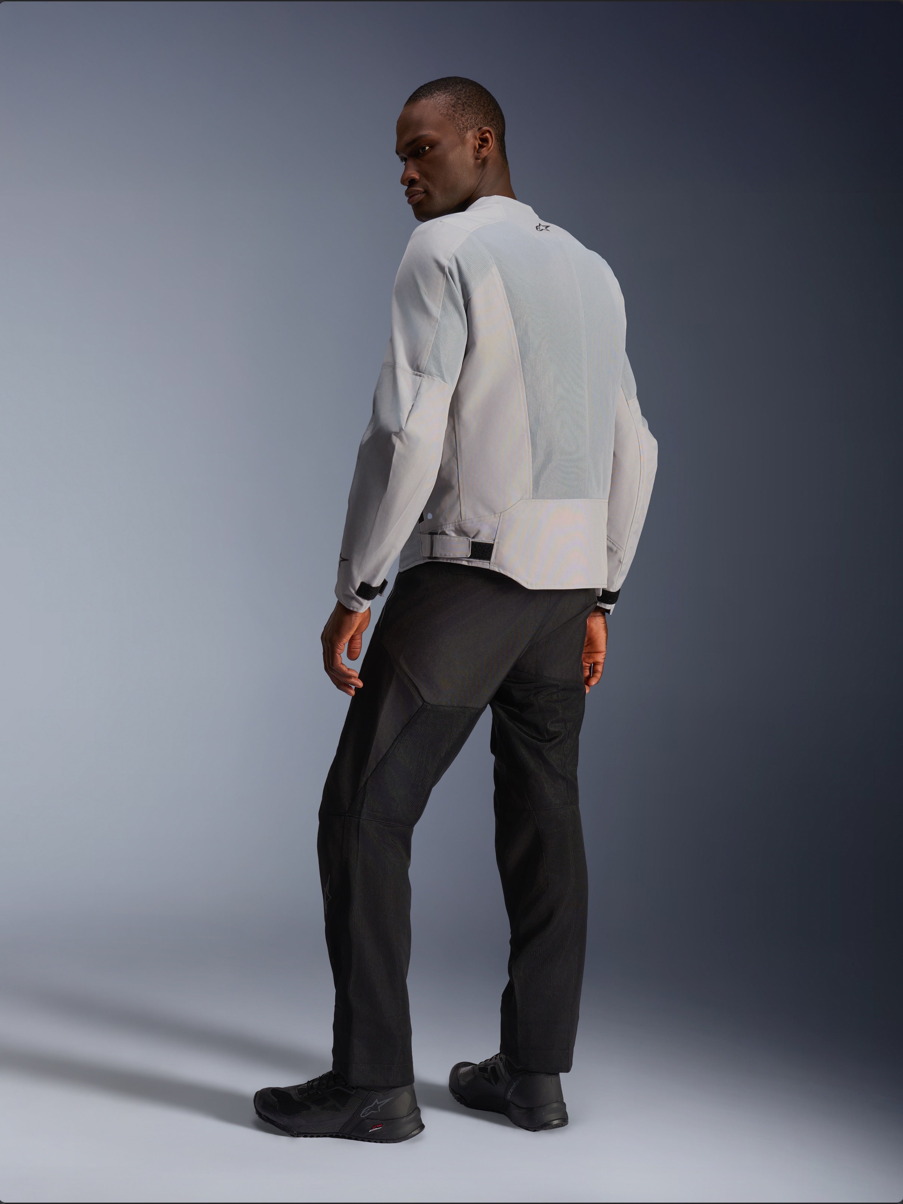 Veste Alpinestars C-1 Air, argent et gris, vue de dos sur un modèle, avec panneaux en maille technique ventilée et logos réfléchissants, portée avec un pantalon et des chaussures noirs, équipement de moto urbain contemporain.