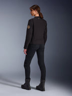 Mannequin portant la veste Alpinestars Stella Grace en noir, vue de dos mettant en évidence l'écusson Oscar vintage en spirale sur la manche et le petit logo étoile sur le col, assortie d'un pantalon de moto noir et de bottes noires pour la conduite urbaine