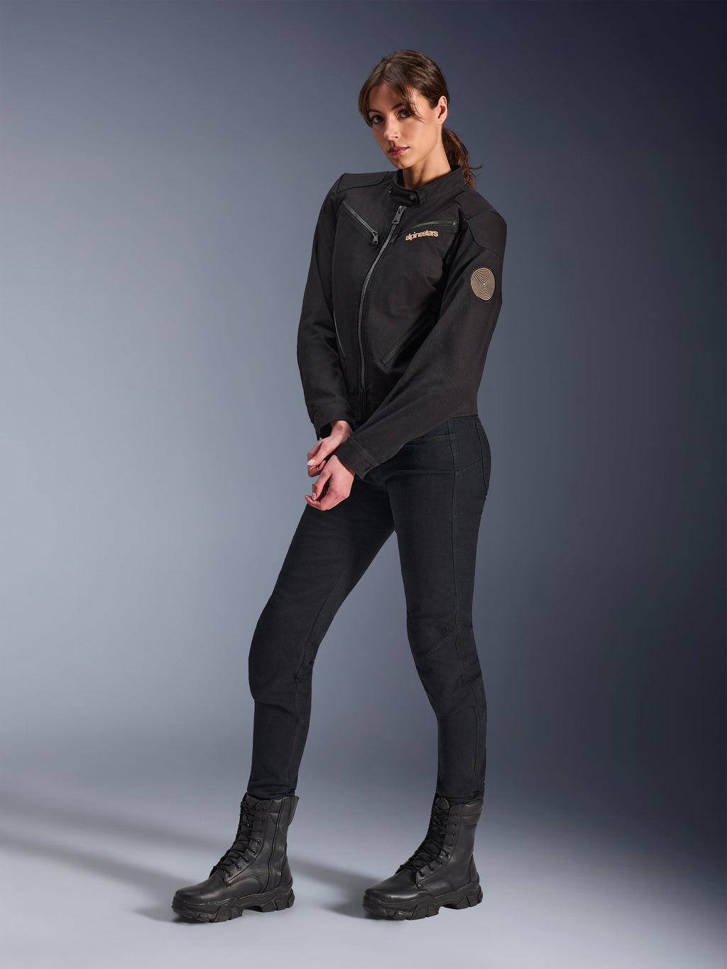 Mannequin femme portant la veste Alpinestars Stella Grace, veste de moto urbaine noire, assortie d'un pantalon de moto et de bottes noirs, design d'inspiration workwear avec un logo brodé bronze sur la poitrine et un écusson circulaire en spirale sur la manche