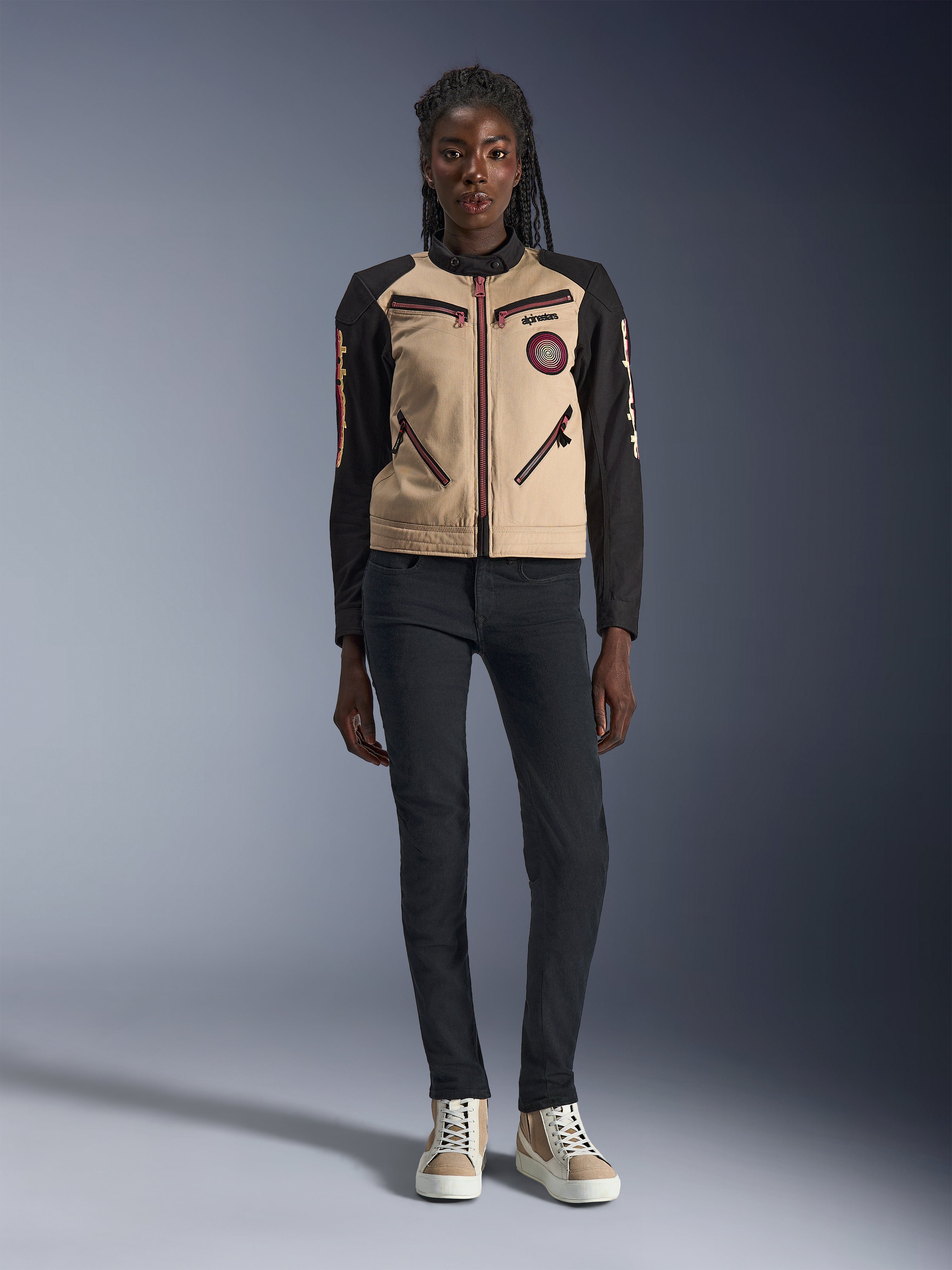 Veste Alpinestars Stella Grace pour femme, veste de moto urbaine, construction textile beige et noire, avec fermetures éclair à l'avant et patch Oscar en spirale brodé, portée avec un pantalon noir et des baskets montantes, équipement de moto d'inspiration workwear contemporaine