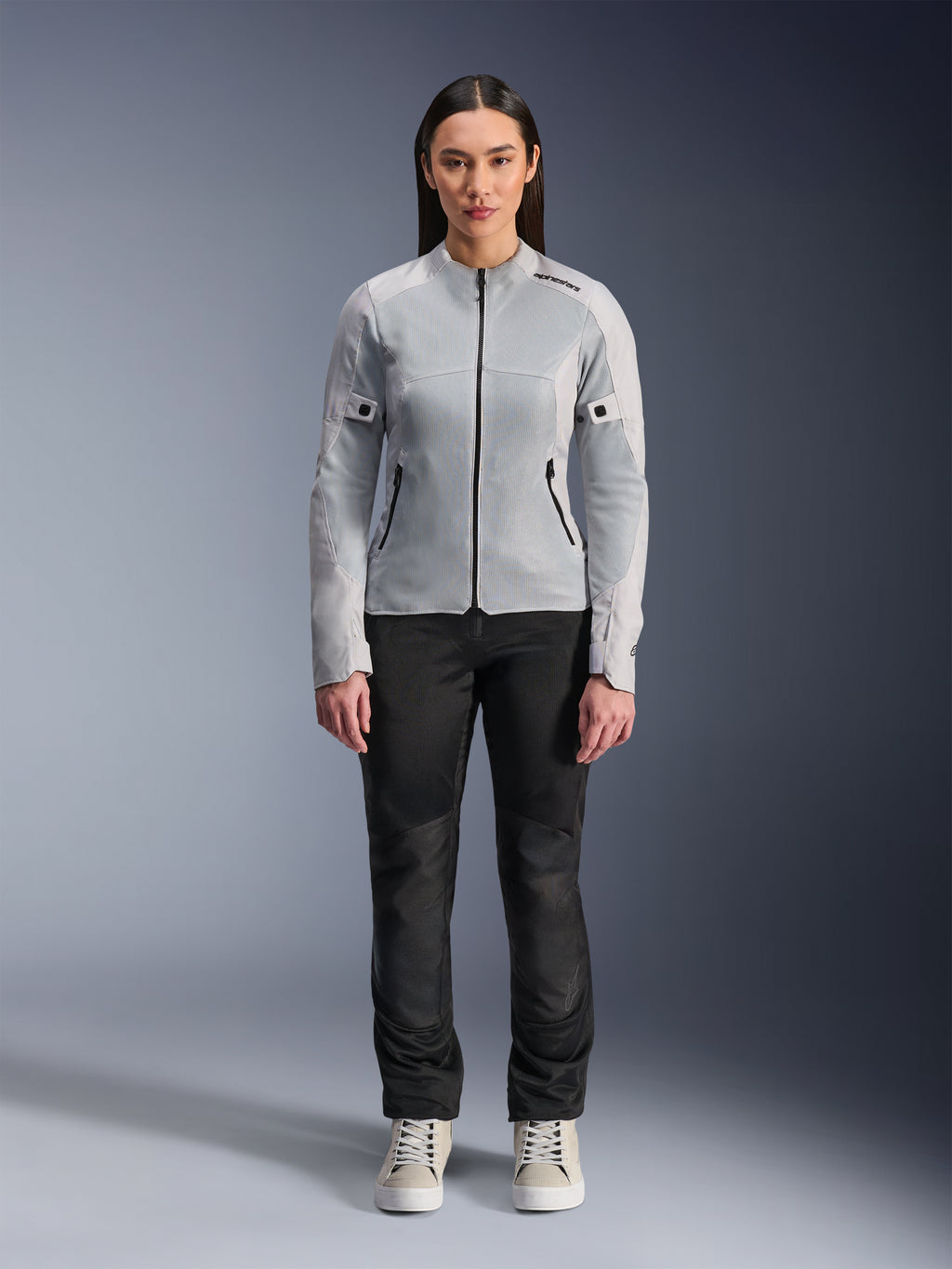 Femme portant la veste Alpinestars Stella C-1 Air en coloris argent, vue de face, veste de moto urbaine en textile avec panneaux en maille technique, associée à un pantalon noir et des baskets blanches, équipement de moto contemporain