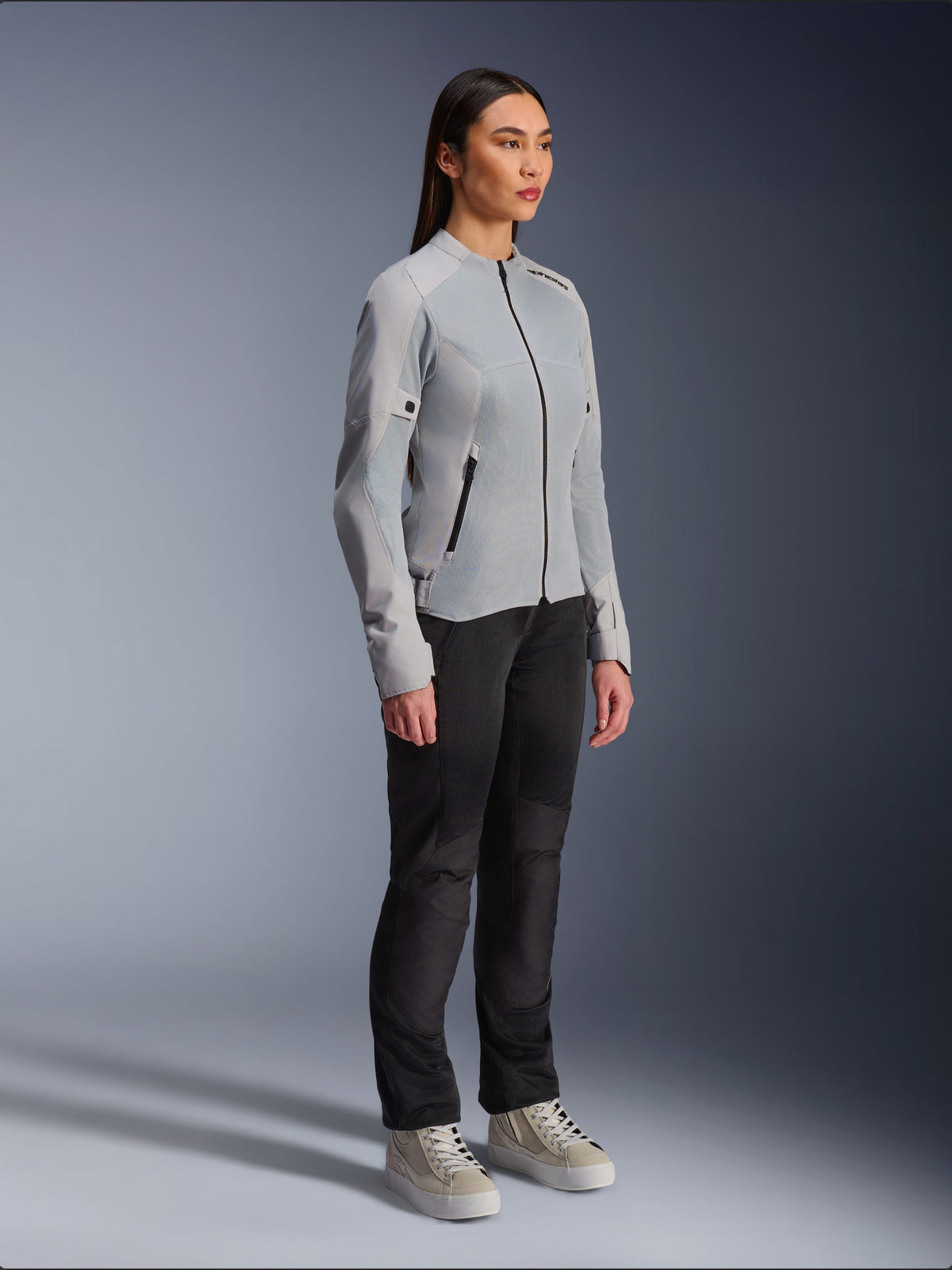 Femme portant la veste Alpinestars Stella C-1 Air en Silver, veste de moto légère en textile et maille, associée à un pantalon de moto noir et des baskets décontractées, esthétique de conduite urbaine épurée.