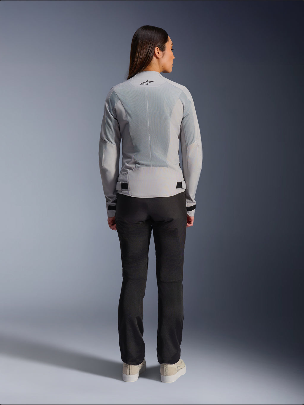 Femme portant la veste Alpinestars Stella C-1 Air couleur argent, vue de dos, dotée de grands panneaux en maille technique pour la ventilation et d'un logo étoile réfléchissant, associée à un pantalon de moto noir et des baskets blanches, équipement de moto urbain contemporain