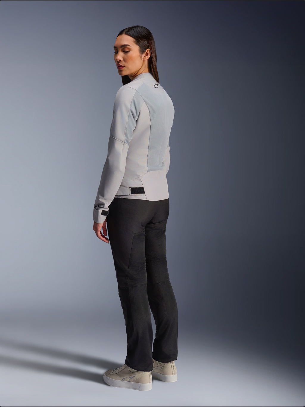 Femme portant la veste Alpinestars Stella C-1 Air en coloris argent, vue de dos et de côté montrant les panneaux en maille technique et le logo réfléchissant sur l'épaule, associée à un pantalon de moto noir et des baskets beiges, esthétique de conduite urbaine contemporaine