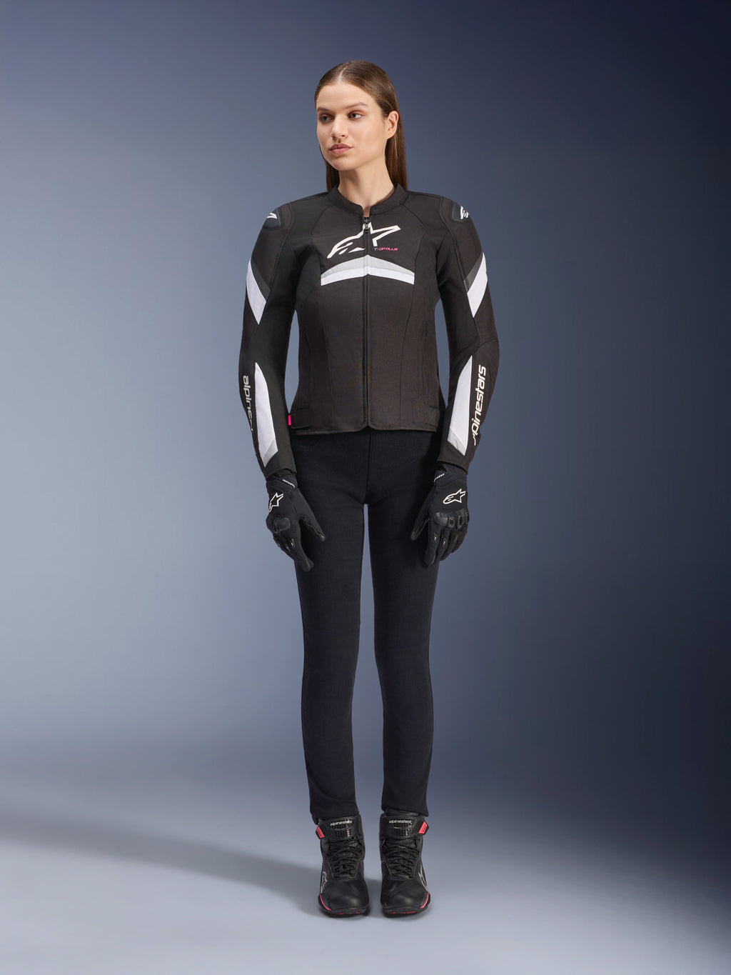Female model wearing the Alpinestars Stella T-GP Plus R V4 Jacket in Black and White, textile sport motorcycle jacket, paired with black riding pants, gloves, and boots, front view showing white Alpinestars logo and sleeve accents.