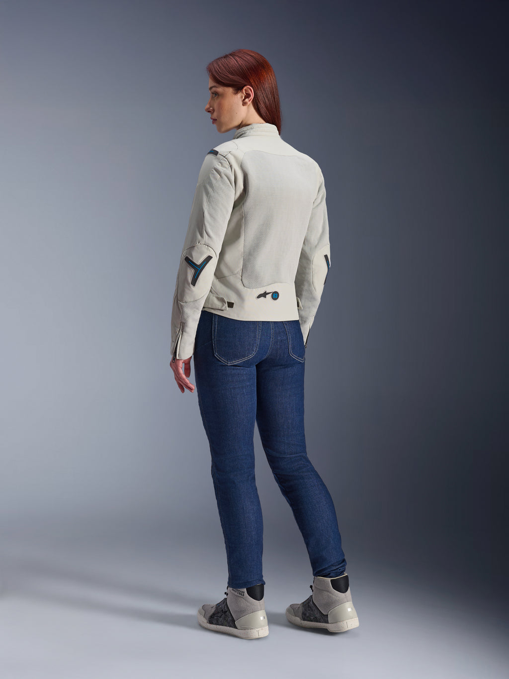 Femme portant la veste Alpinestars Stella Circal-1 Air en Metal Gray et Methyl Blue, vue arrière montrant les panneaux de ventilation en mesh et les accents bleu néon, associée à un jean de moto bleu foncé et des baskets grises, photo lifestyle moto urbaine.