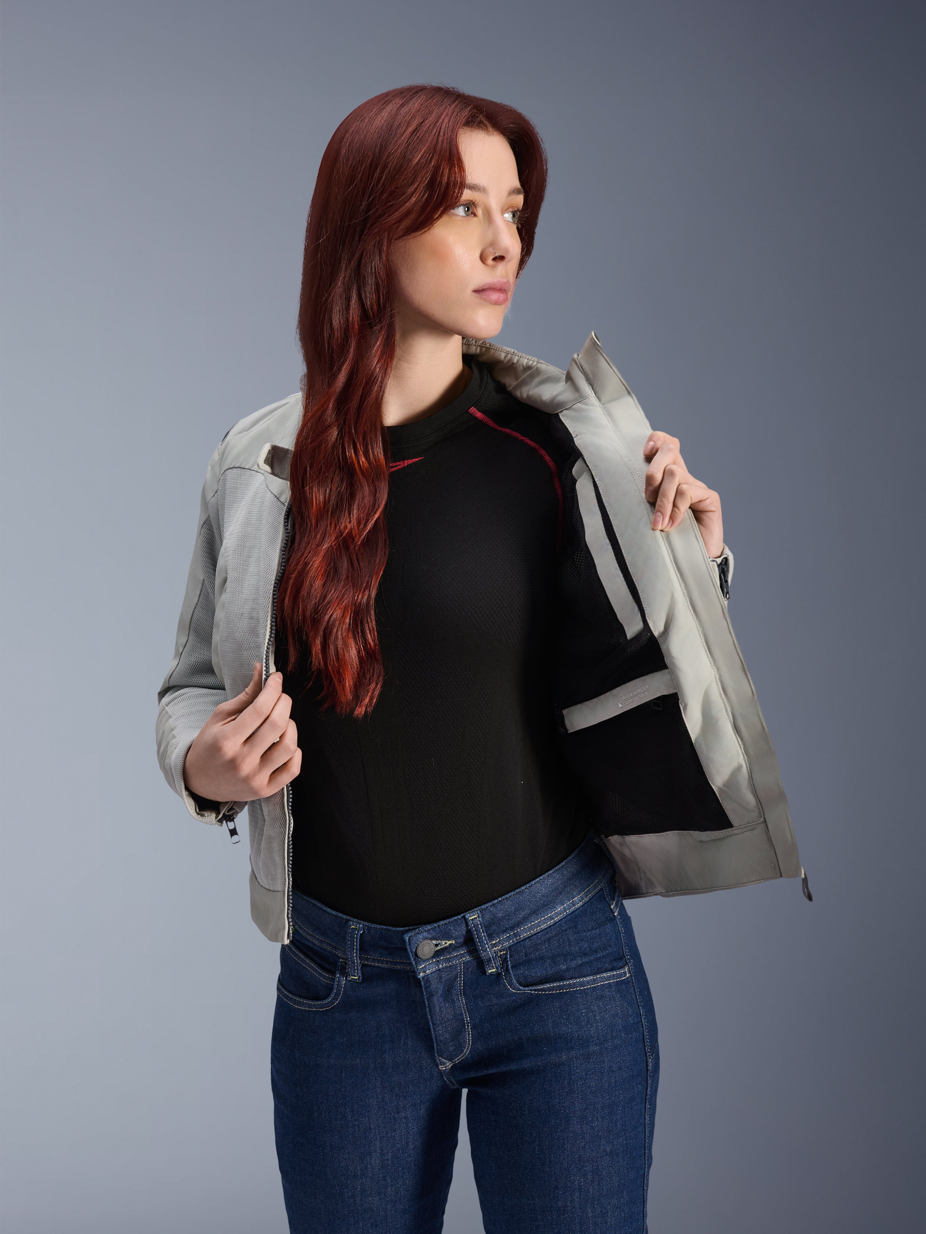 Veste Alpinestars Stella Circal-1 Air en Gris Métal, modèle féminin ouvrant la veste pour révéler la doublure intérieure en mesh et la poche, portée avec un jean bleu et un haut noir, équipement de moto urbain contemporain.