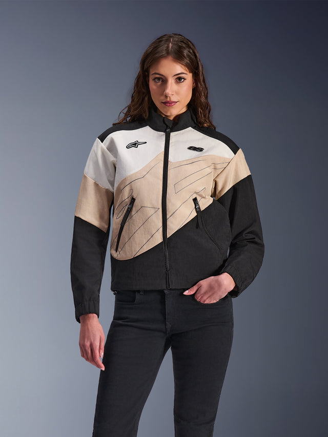 Veste Alpinestars Stella Moshell, veste de moto urbaine pour femme, design asymétrique à blocs de couleurs en noir, sable et blanc cassé, dotée d'une fermeture éclair frontale et de poches zippées, portée avec un pantalon noir pour un look streetwear contemporain.