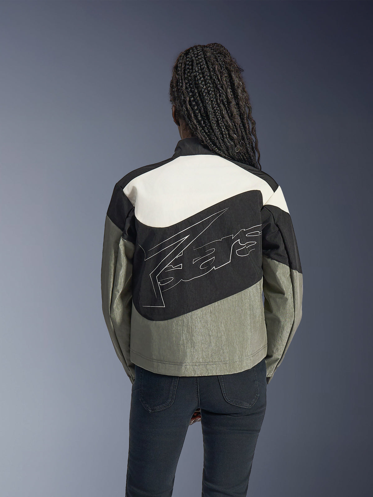 Woman from behind wearing the Alpinestars Stella Moshell Jacket, Black Gray and Off White urban motorcycle jacket, featuring a large white outlined logo on the back, paired with black pants, contemporary streetwear-inspired riding gear.