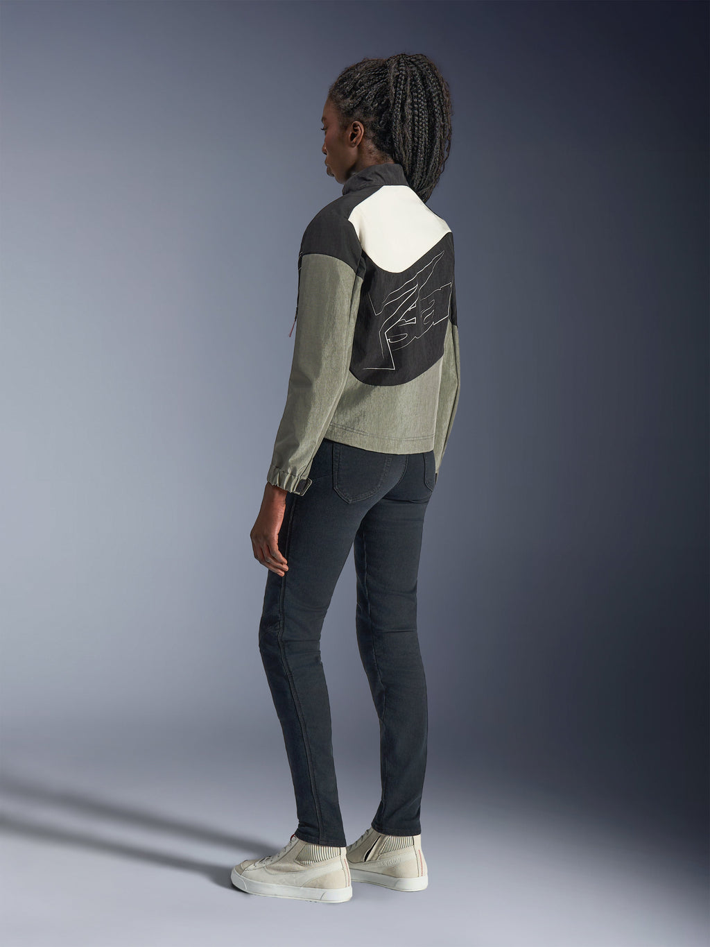 Woman wearing Alpinestars Stella Moshell Jacket, Urban Motorcycle Jacket, Black, Gray, and Off-White colorway, rear view featuring asymmetrical panel design and large back graphic, styled with black slim-fit pants and off-white sneakers, contemporary urban motorcycle gear