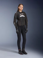 Motarde portant la veste Alpinestars Stella T-GP Plus R V4 Airflow, noir et blanc, dotée de panneaux en mesh pour la ventilation et de logos blancs, associée à un pantalon noir, des gants noirs et des bottes noires avec des accents roses, équipement de moto de sport pour femme