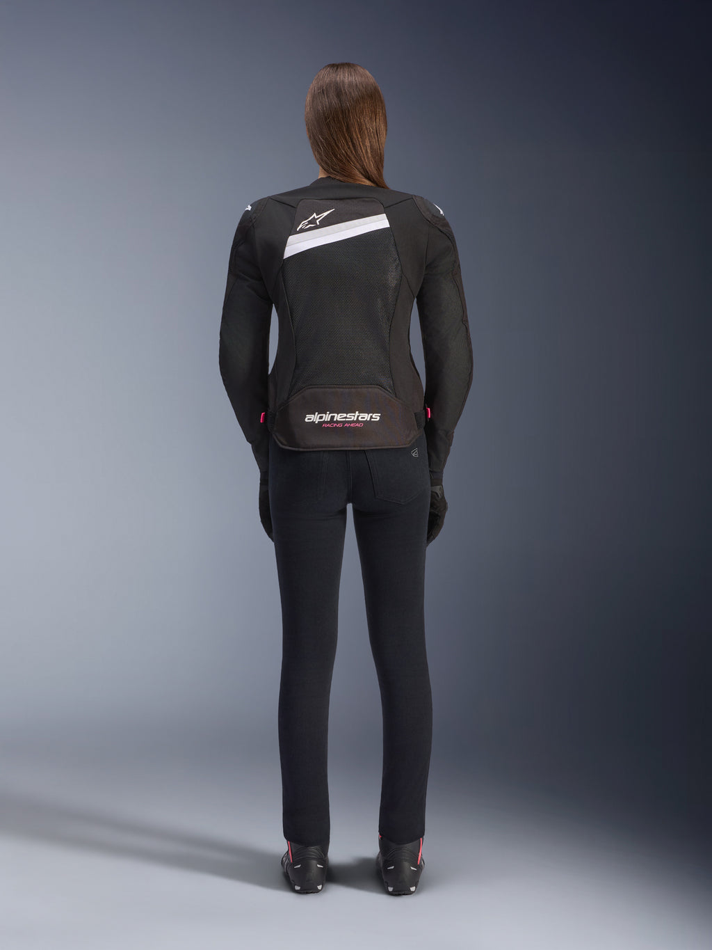 Veste Alpinestars Stella T-GP Plus R V4 Airflow, noir et blanc, vue de dos montrant les larges panneaux de ventilation en mesh et le logo Alpinestars, portée par une motarde avec un pantalon de moto noir coupe slim et des bottes, équipement textile de sport pour femme