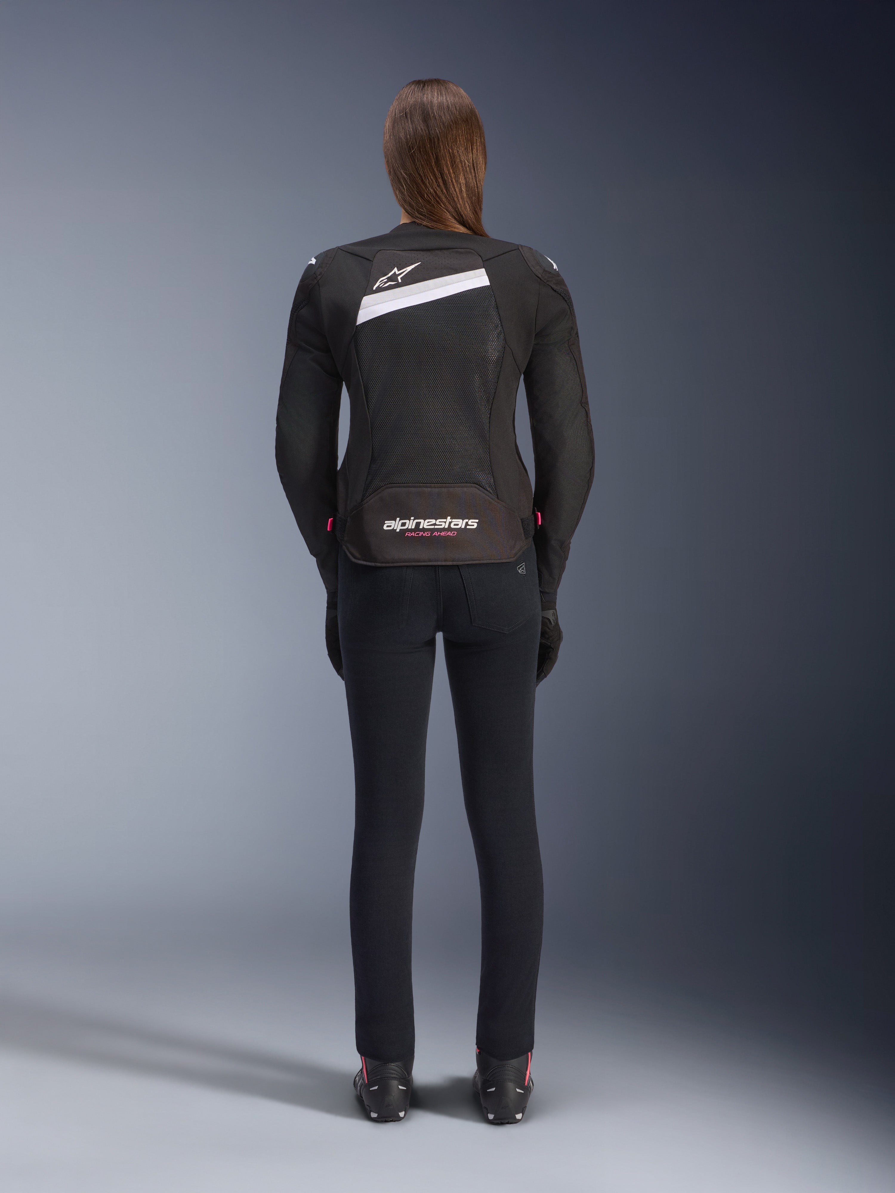 Veste Alpinestars Stella T-GP Plus R V4 Airflow, noir et blanc, vue de dos montrant les larges panneaux de ventilation en mesh et le logo Alpinestars, portée par une motarde avec un pantalon de moto noir coupe slim et des bottes, équipement textile de sport pour femme