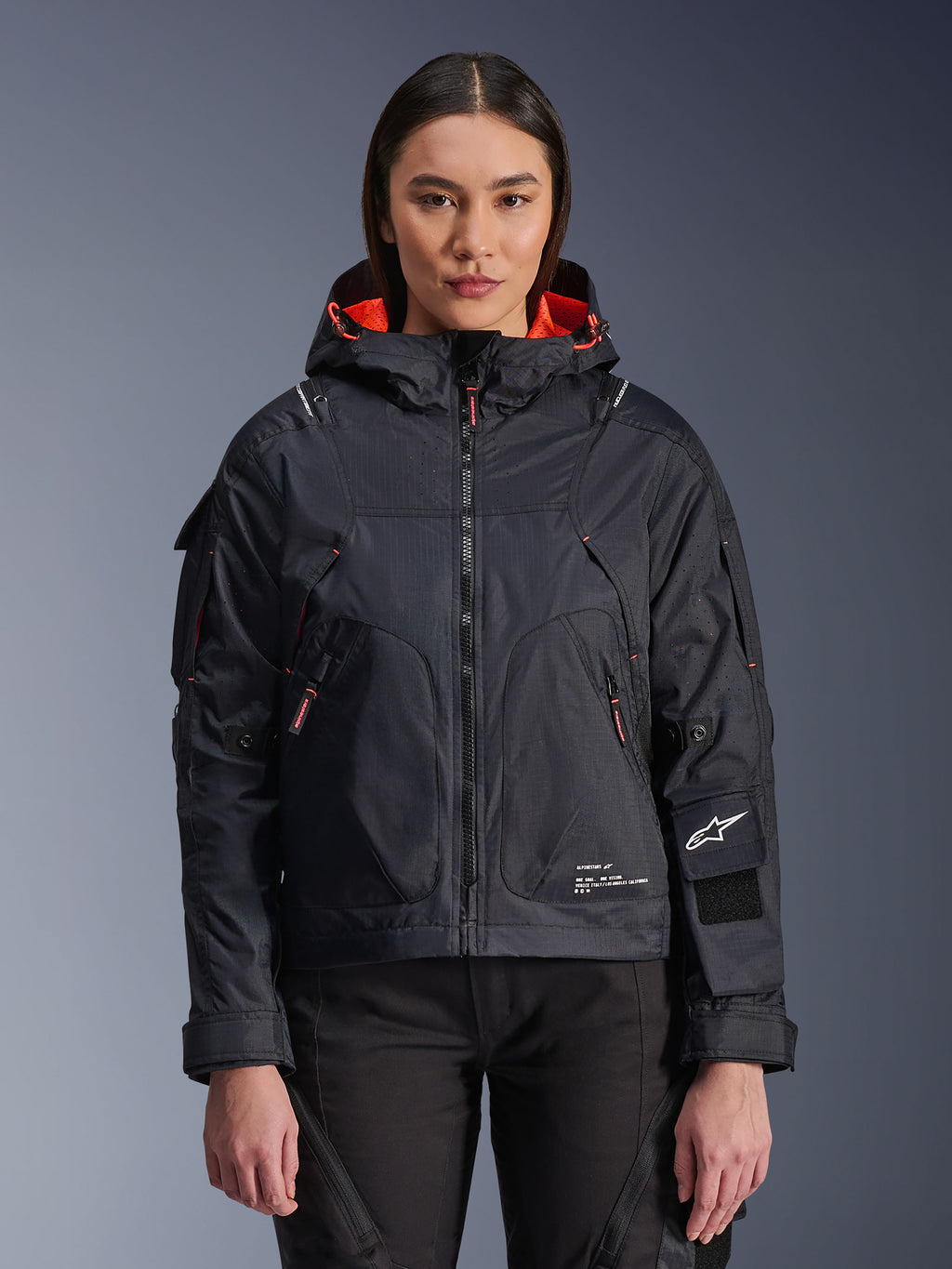 Alpinestars Molly Air Jacket, veste de moto urbaine pour femme, noire avec doublure intérieure corail fluo, portée par un mannequin femme, design à capuche en textile technique, poches zippées et logo Alpinestars sur la manche