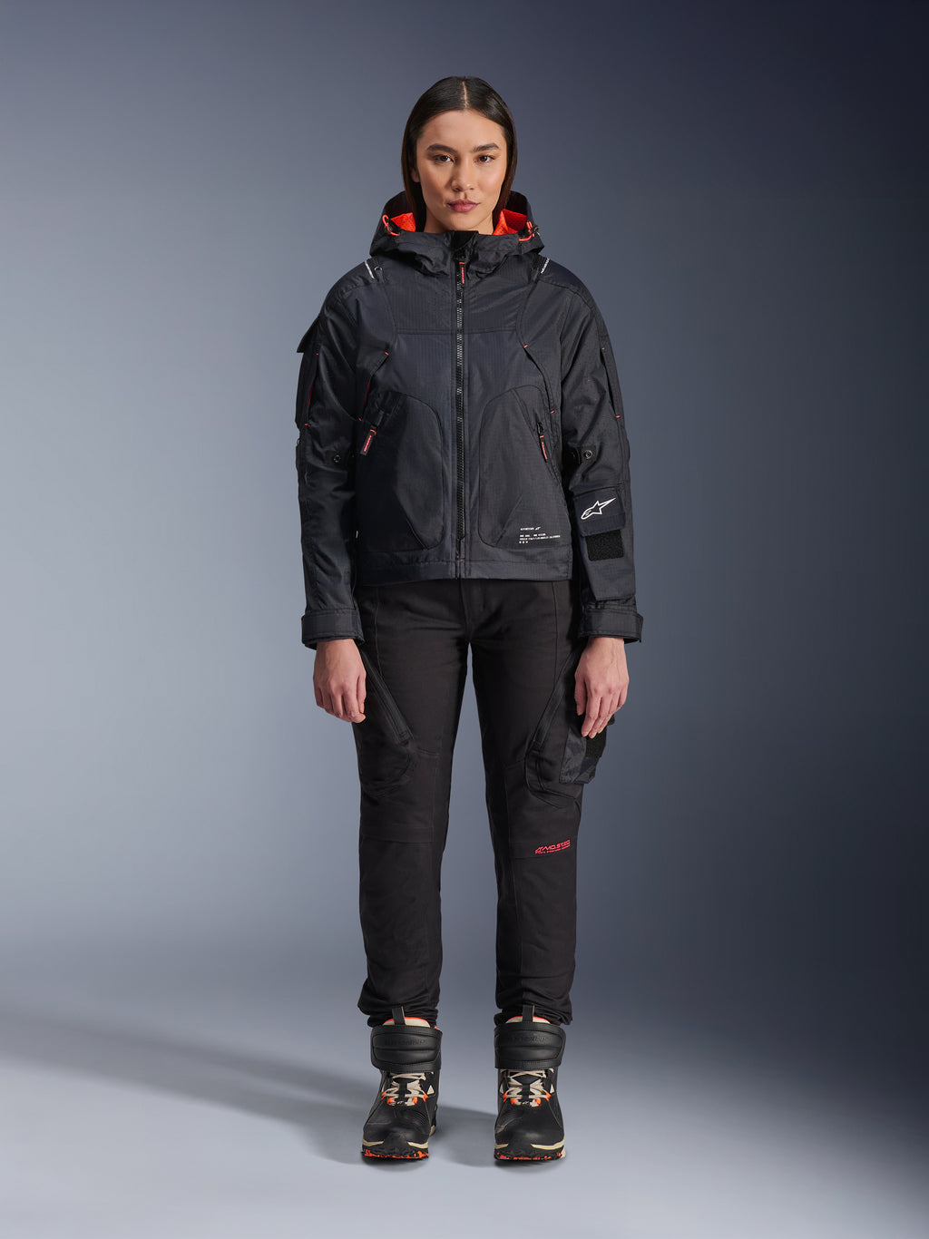 Mannequin femme portant la veste Alpinestars Molly Air en Noir et Corail Fluo, design d'inspiration utilitaire avec capuche et doublure corail, associée à un pantalon de moto noir et des bottes de moto, équipement de moto urbain contemporain