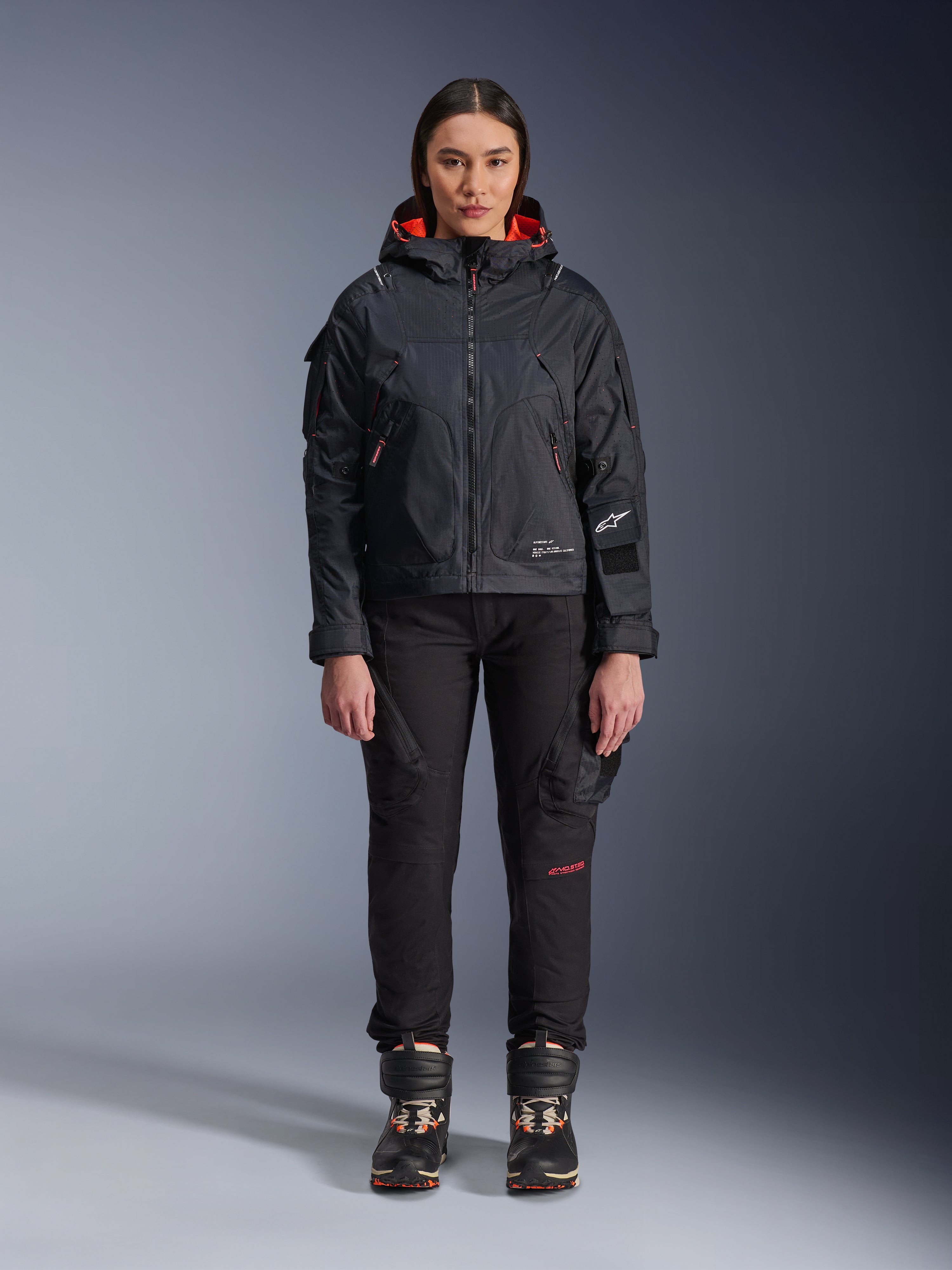 Mannequin femme portant la veste Alpinestars Molly Air en Noir et Corail Fluo, design d'inspiration utilitaire avec capuche et doublure corail, associée à un pantalon de moto noir et des bottes de moto, équipement de moto urbain contemporain