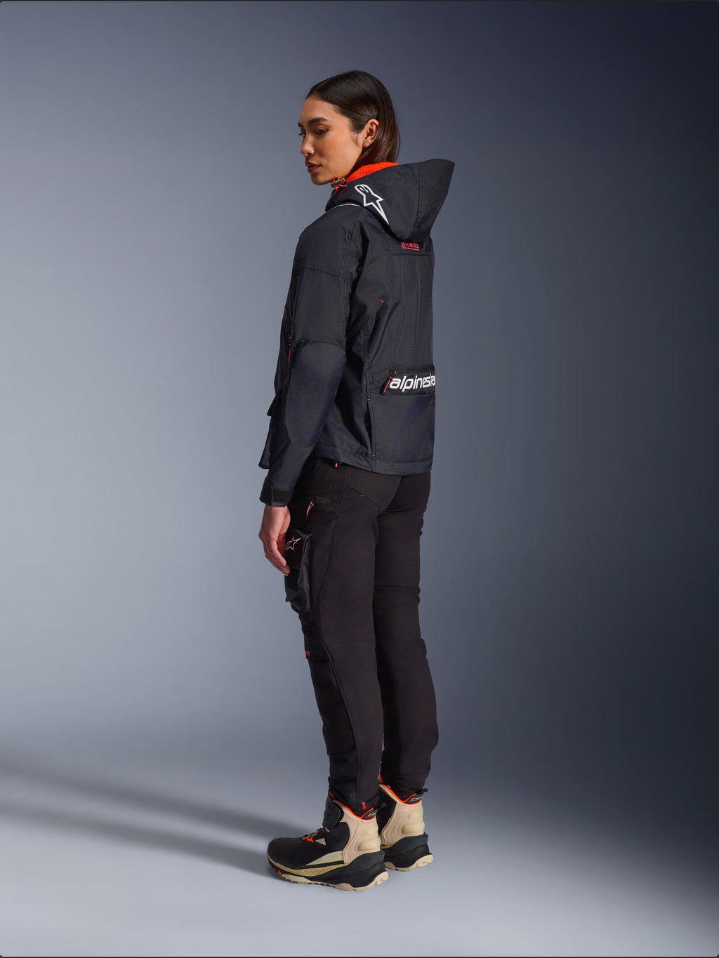 Pantalon femme Alpinestars Actea, noir, pantalon de moto moderne avec coupe fuselée et poche cargo sur la cuisse, vue de profil arrière sur un mannequin portant une veste à capuche sombre et des bottes de moto, style urbain