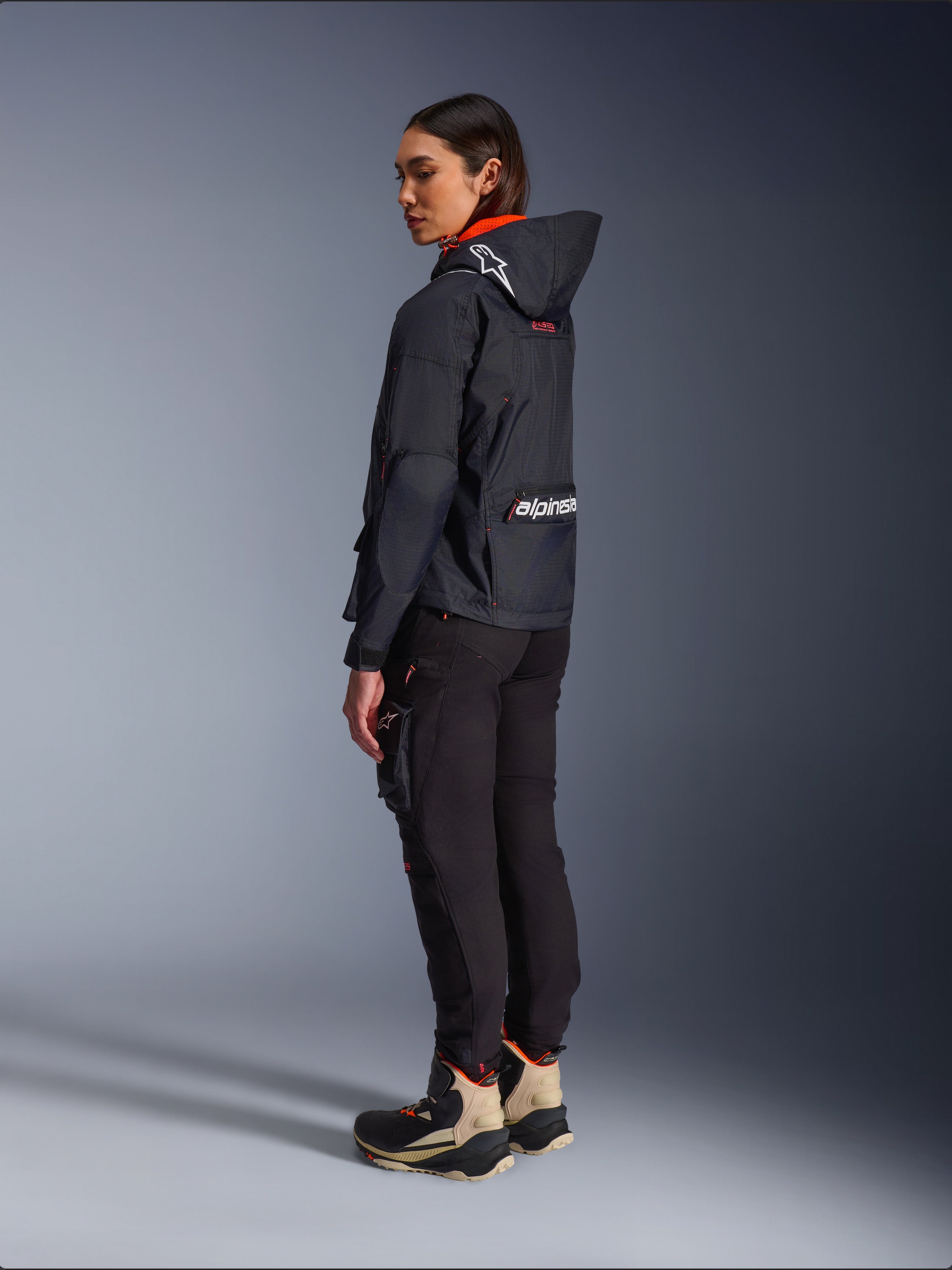 Pantalon femme Alpinestars Actea, noir, pantalon de moto moderne avec coupe fuselée et poche cargo sur la cuisse, vue de profil arrière sur un mannequin portant une veste à capuche sombre et des bottes de moto, style urbain