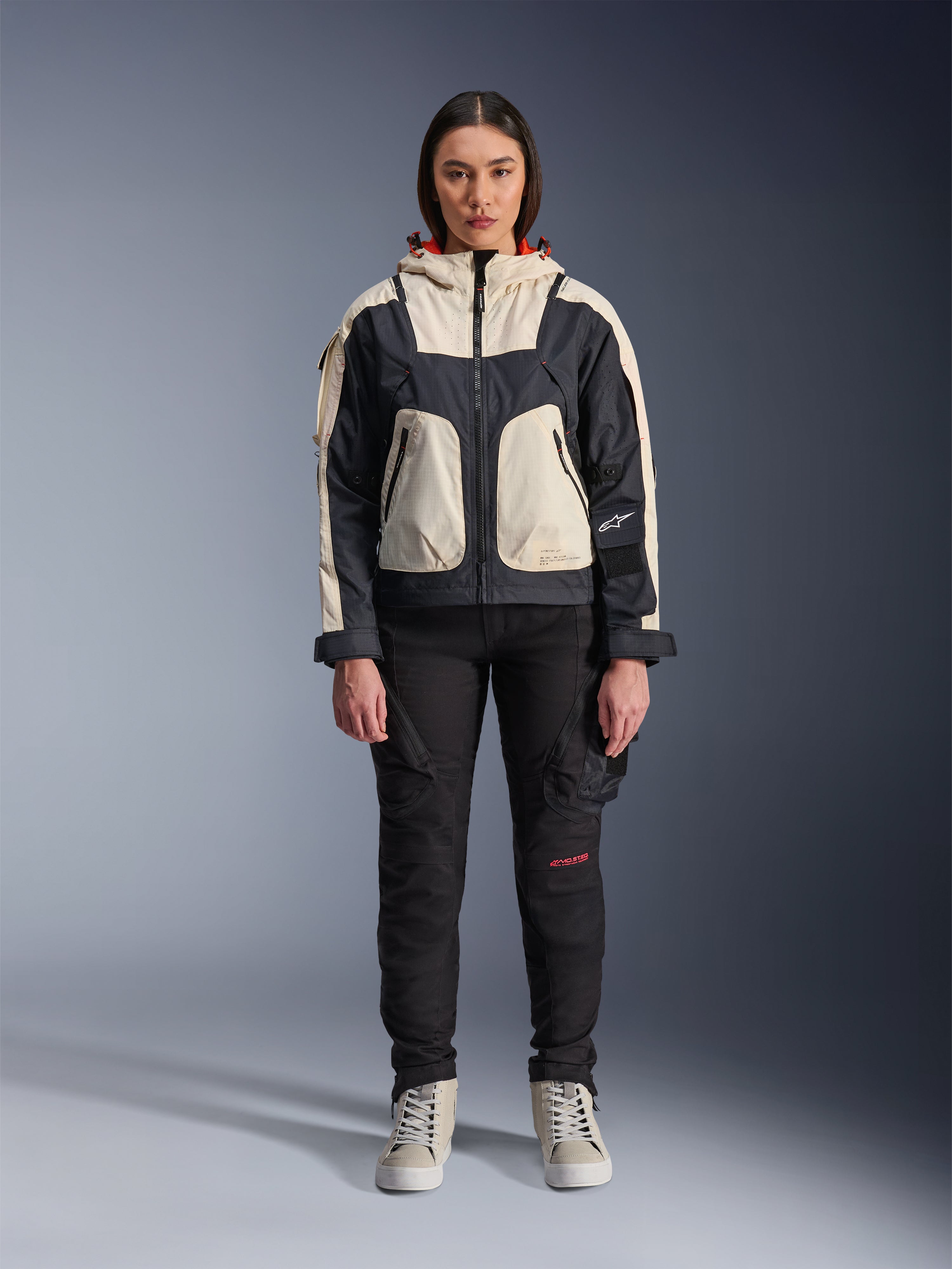 Mannequin femme portant la veste Alpinestars Molly Air en Black Almond, design color-block avec empiècements crème et noirs et capuche doublée de rouge, pantalon de moto noir et baskets crème, équipement de moto urbain contemporain, photo de studio