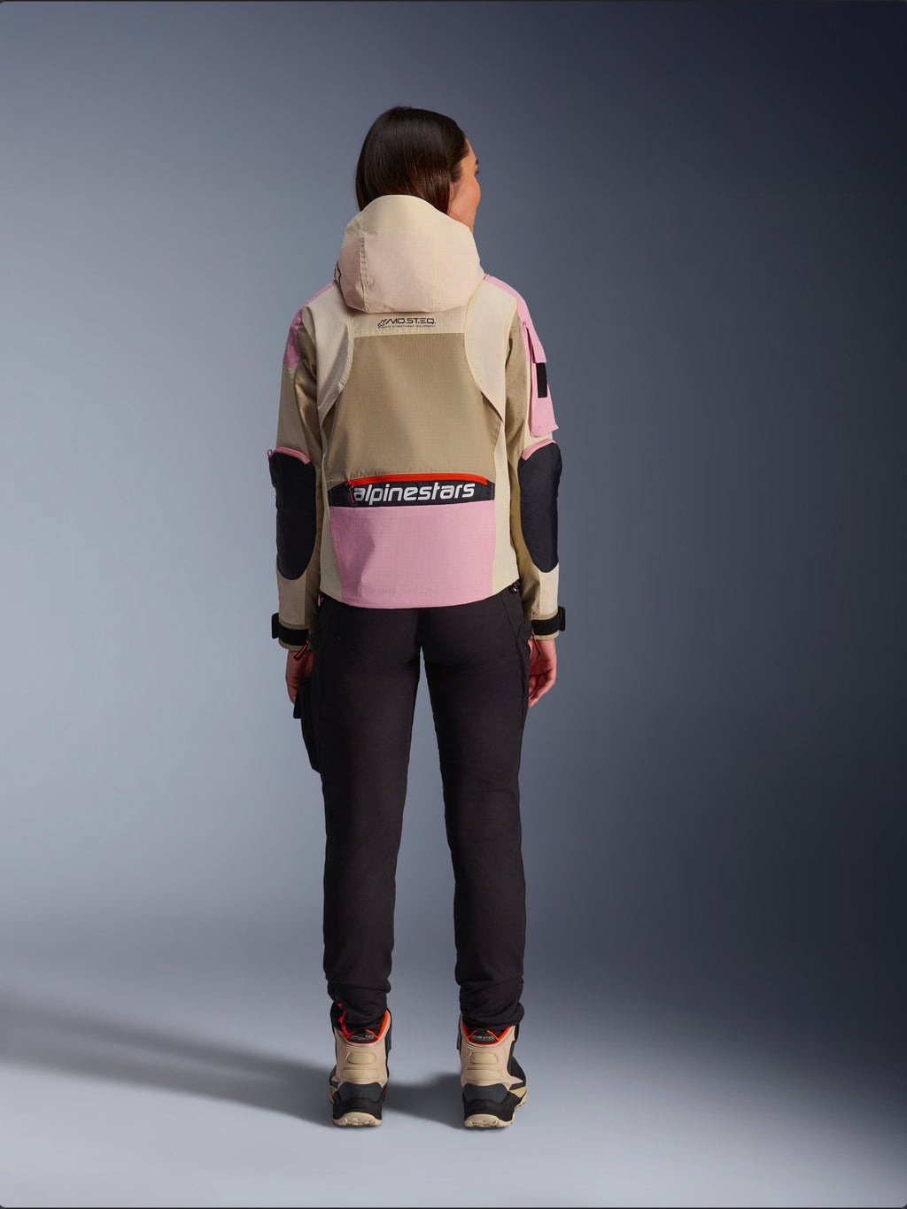 Motarde vue de dos portant la veste Alpinestars Molly Air en coloris Sand Pink Almond, dotée d'une capuche beige et d'empiècements roses avec des accents noirs, associée à un pantalon de moto noir et des bottes beiges, vêtement de moto urbain contemporain