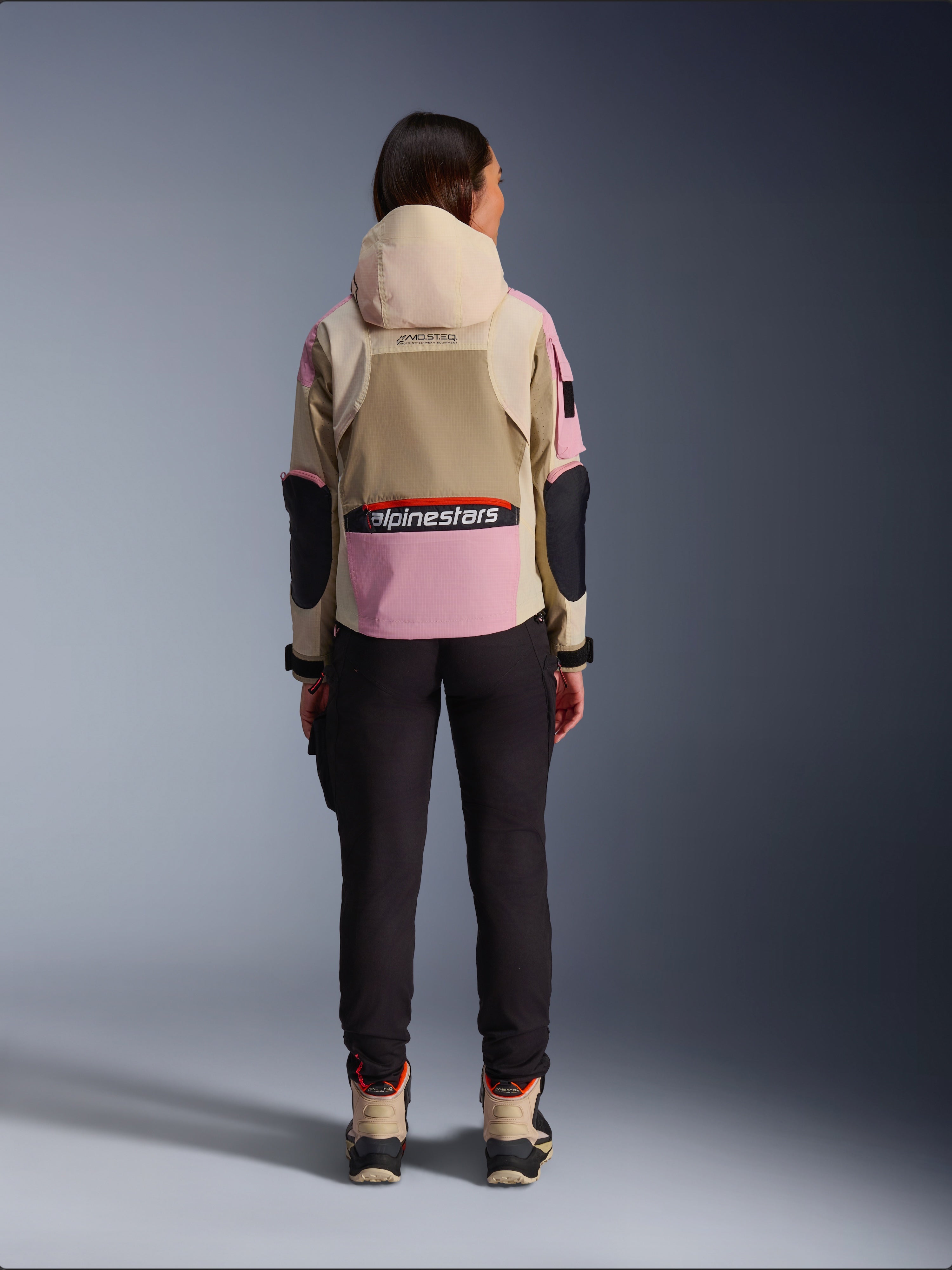 Motarde vue de dos portant la veste Alpinestars Molly Air en coloris Sand Pink Almond, dotée d'une capuche beige et d'empiècements roses avec des accents noirs, associée à un pantalon de moto noir et des bottes beiges, vêtement de moto urbain contemporain