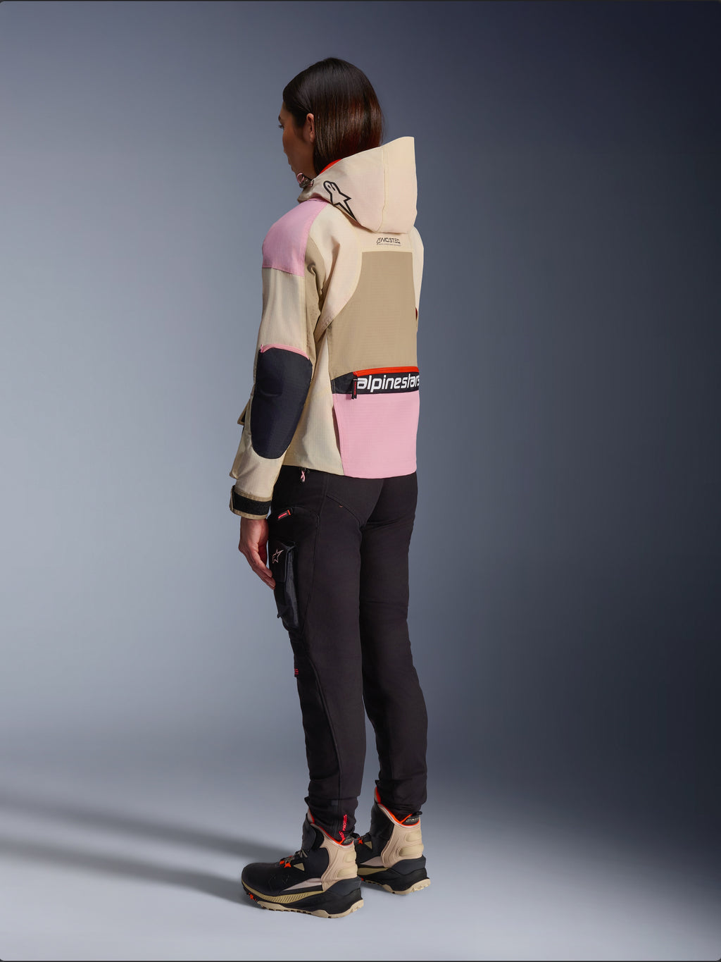 Motarde portant la veste Alpinestars Molly Air en Sand Pink Almond, design beige et rose multi-tons, vue arrière latérale avec pantalon de moto noir et bottes techniques assorties, style moto urbain avec capuche.