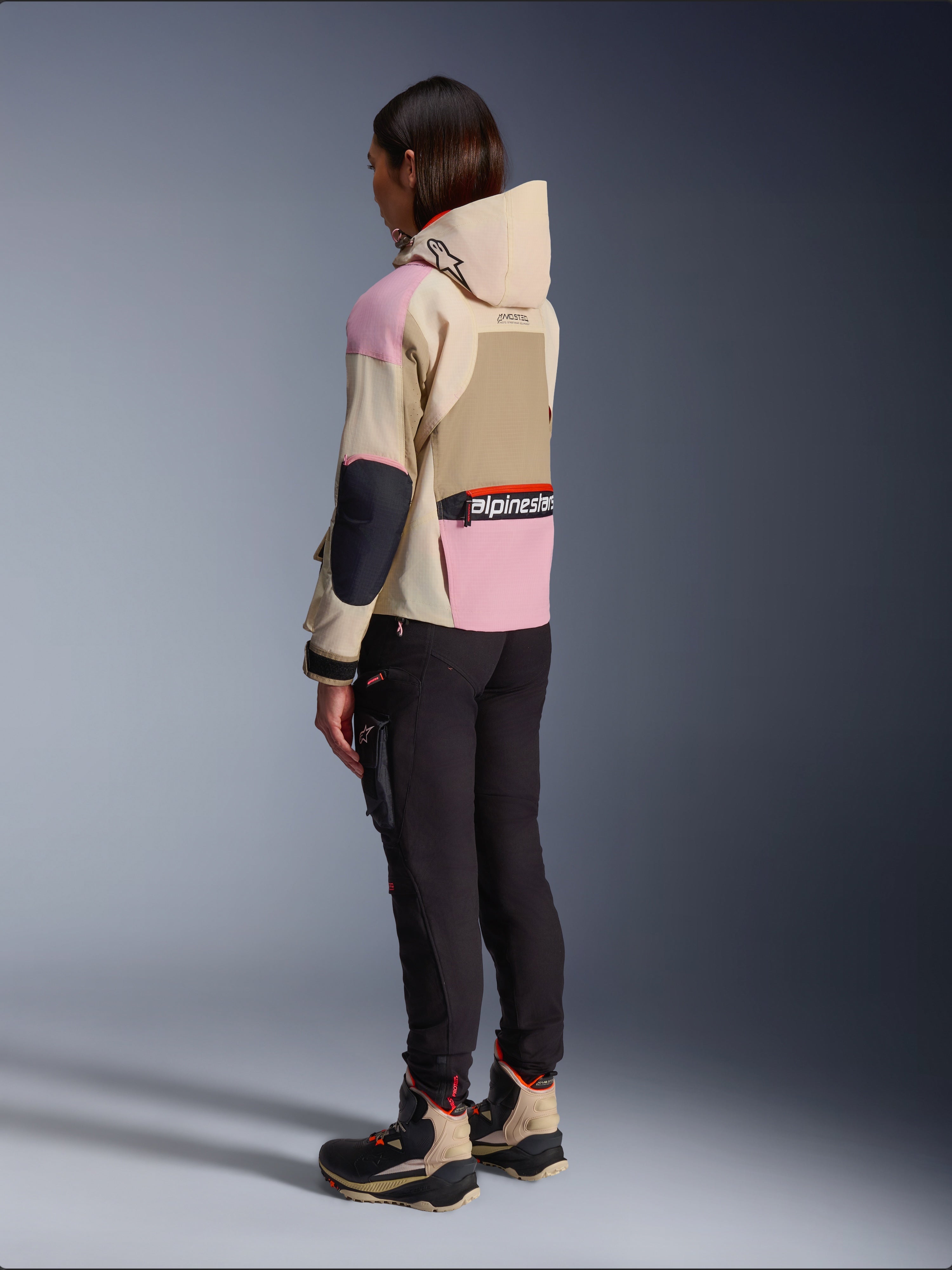 Motarde portant la veste Alpinestars Molly Air en Sand Pink Almond, design beige et rose multi-tons, vue arrière latérale avec pantalon de moto noir et bottes techniques assorties, style moto urbain avec capuche.