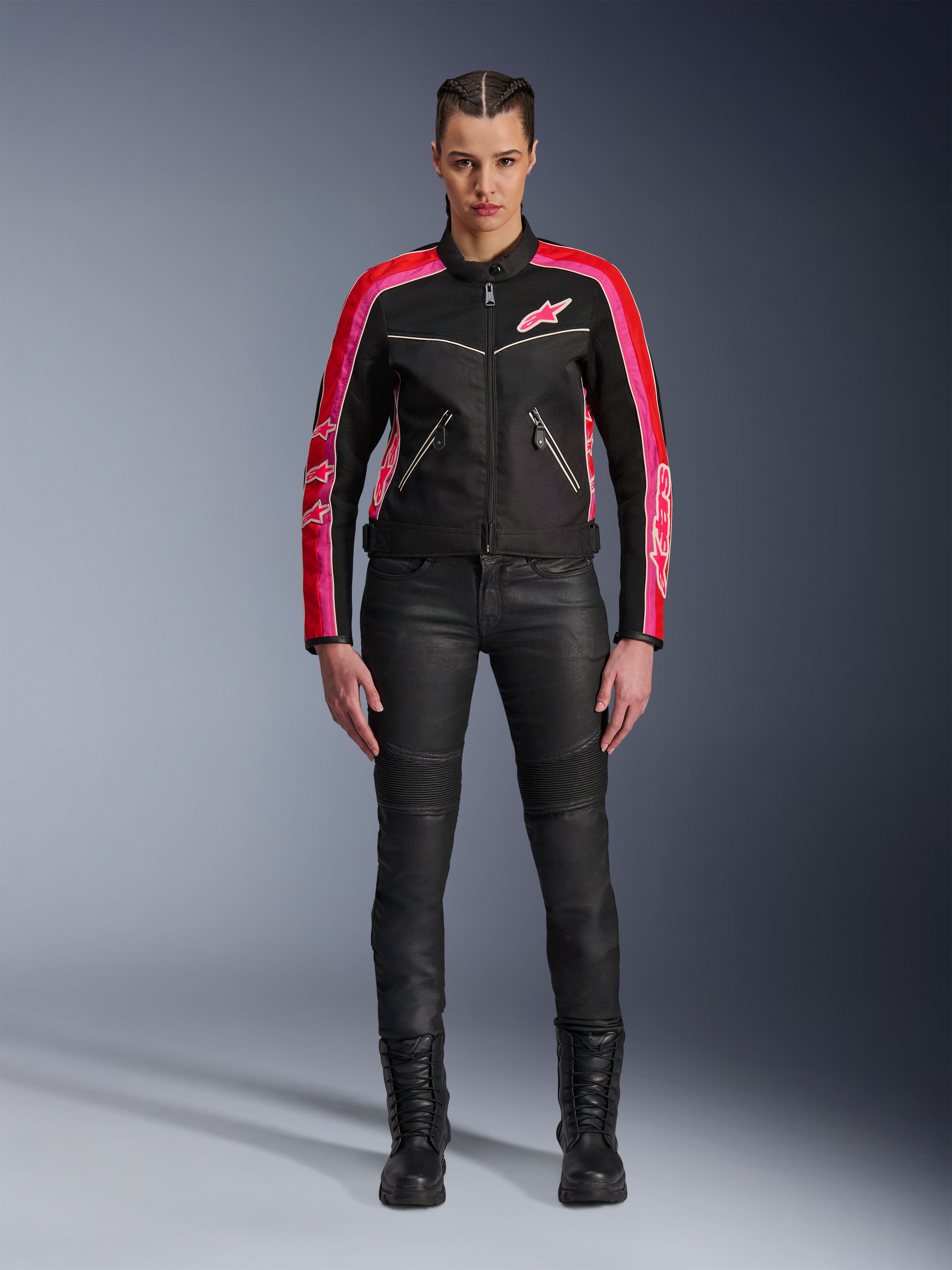 Motarde portant la veste Alpinestars Stella T-Dyno Air, noire avec des accents rouge vif et fuchsia, associée à un pantalon de moto noir et des bottes à lacets, avec panneaux en maille 3D, coupe ajustée et logos d'étoiles roses, tenue d'équipement de moto urbaine