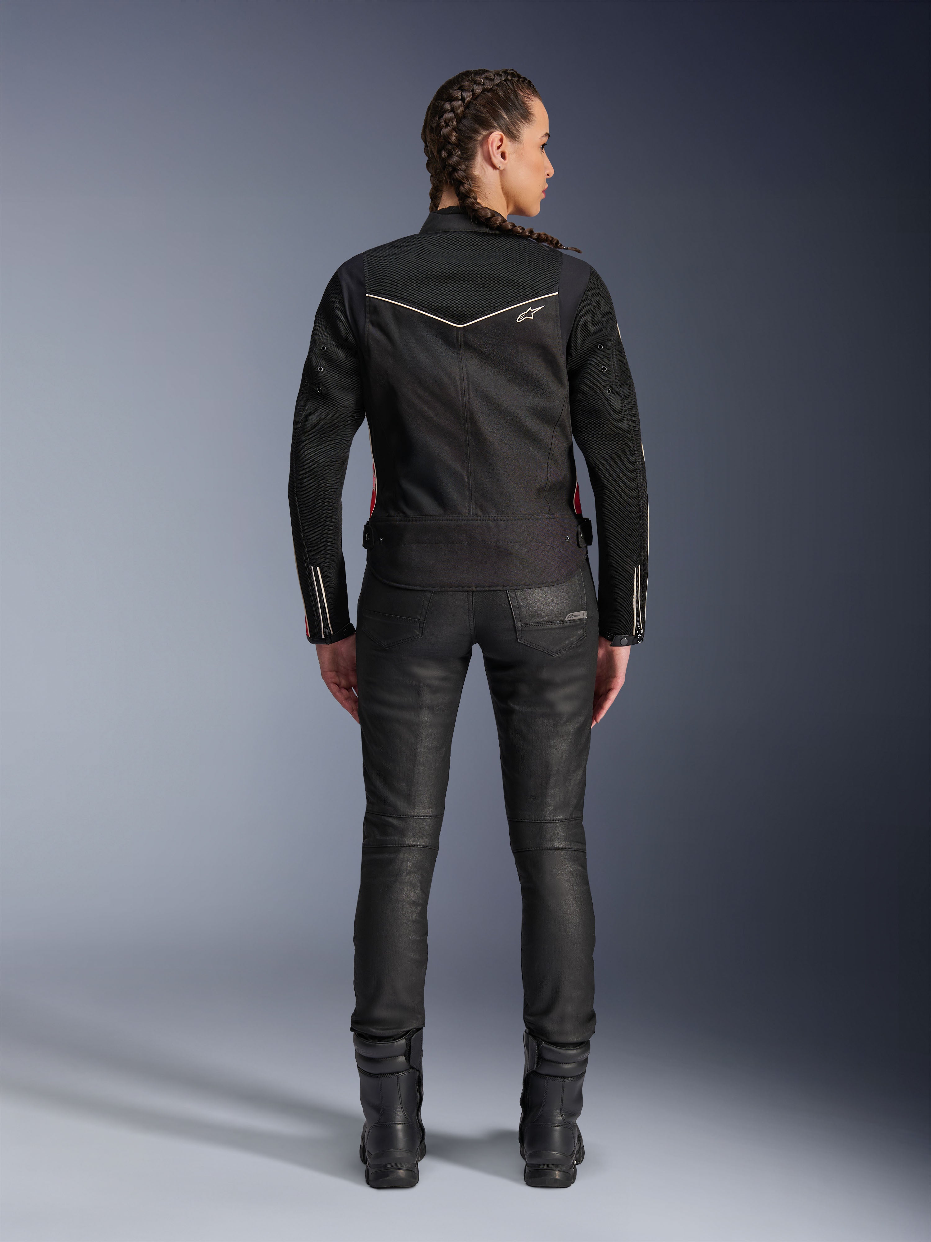 Veste Alpinestars Stella T-Dyno Air, vue arrière, veste de moto noire pour femme avec liseré blanc et petit logo étoile, portée avec un pantalon technique noir et des bottes de moto, coupe biker élégante pour la conduite urbaine