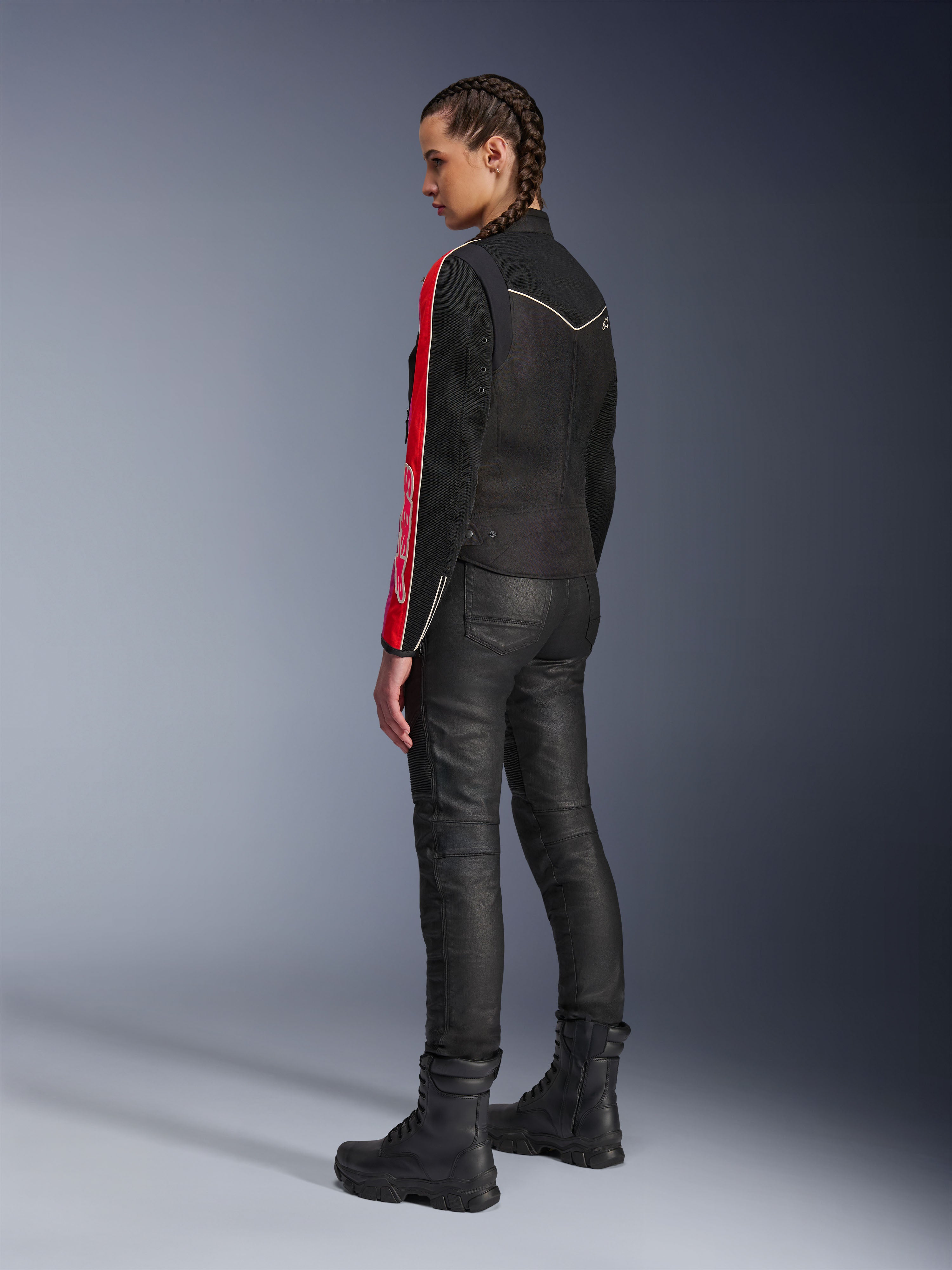 Femme portant la veste Alpinestars Stella T-Dyno Air en Noir Rouge Vif Fuchsia, associée à un pantalon de moto noir et des bottes noires, vue de profil mettant en valeur les graphismes rouges et fuchsia sur les manches et la coupe biker ajustée.