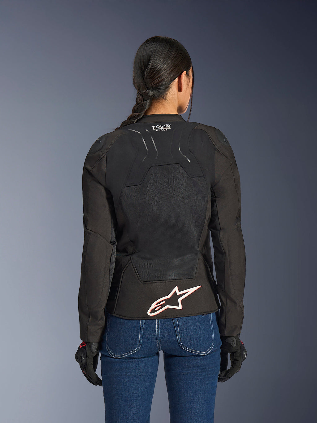 Veste Alpinestars Stella SMX Air, vue de dos sur un modèle féminin, textile noir avec panneaux en maille haute ventilation, marquage 'TECH-AIR READY' sur le haut du dos, grand logo Alpinestars blanc avec contour rouge sur le bas du dos, associée à un jean en denim bleu et des gants de moto noirs