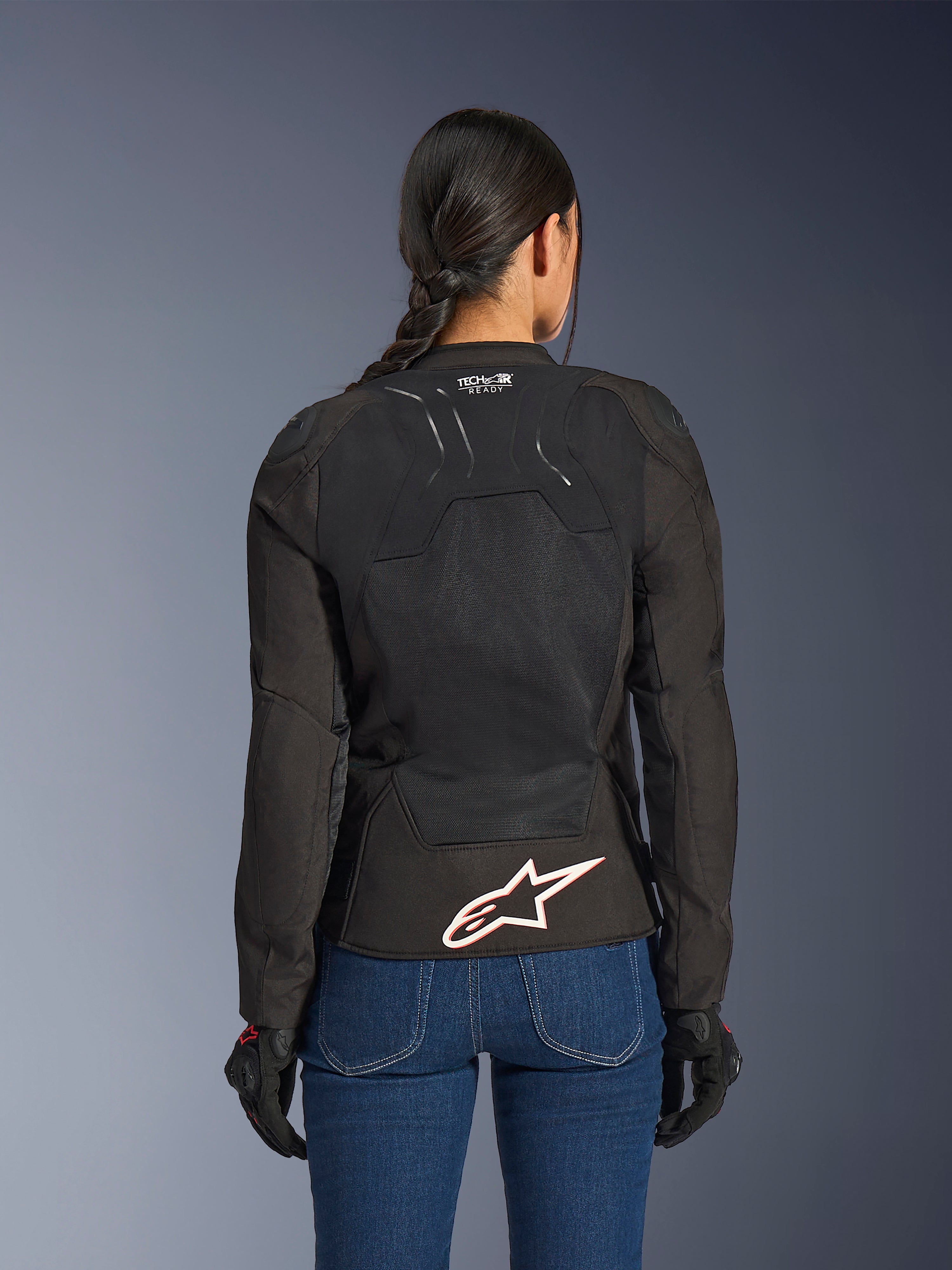 Veste Alpinestars Stella SMX Air, vue de dos sur un modèle féminin, textile noir avec panneaux en maille haute ventilation, marquage 'TECH-AIR READY' sur le haut du dos, grand logo Alpinestars blanc avec contour rouge sur le bas du dos, associée à un jean en denim bleu et des gants de moto noirs