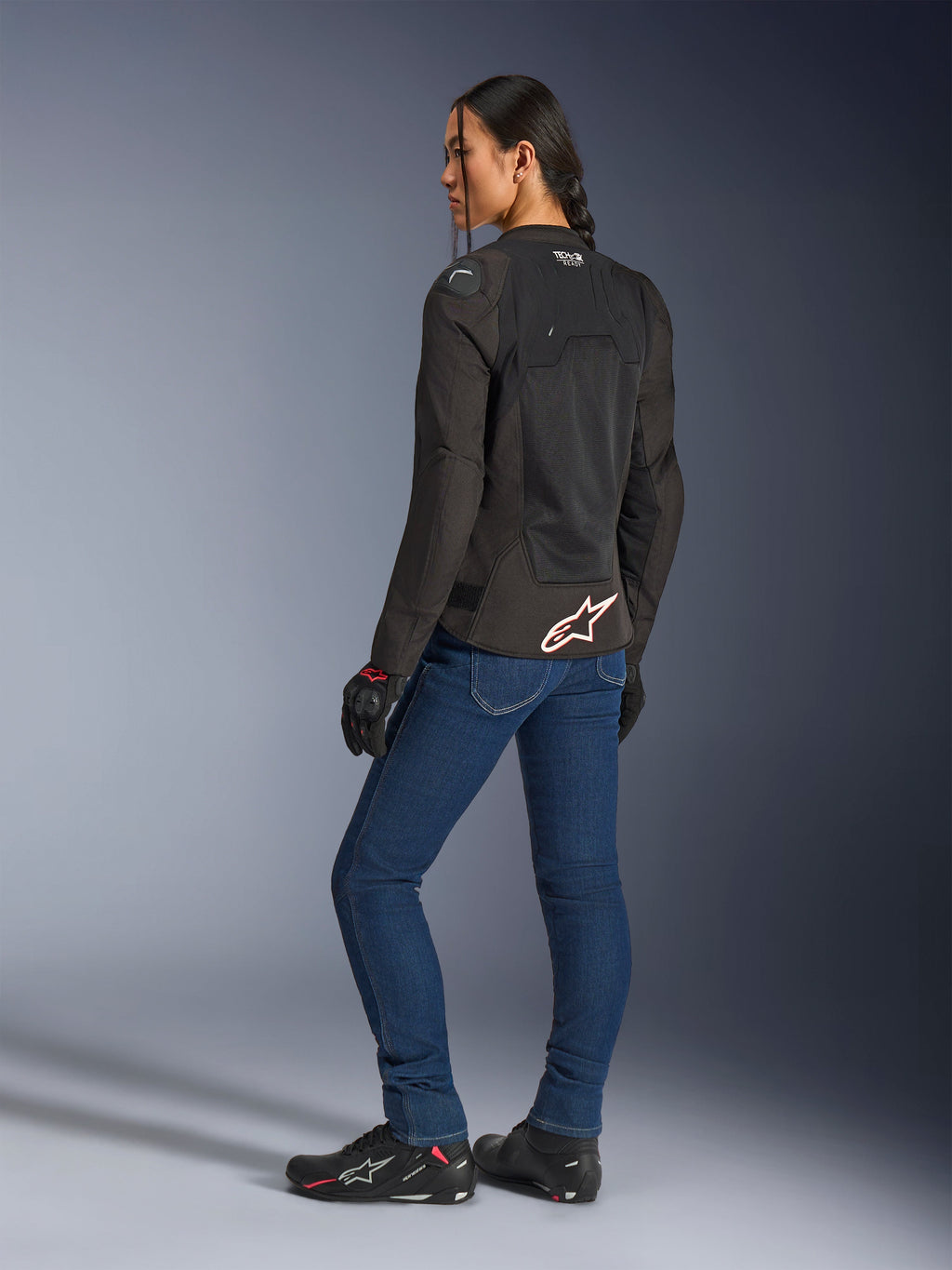 Veste Alpinestars Stella SMX Air, noire avec logo blanc et rouge, portée par une motarde vue de trois-quarts arrière, associée à un jean de moto bleu et des chaussures de moto noires, dotée d'une construction technique en mesh pour la conduite sportive par temps chaud