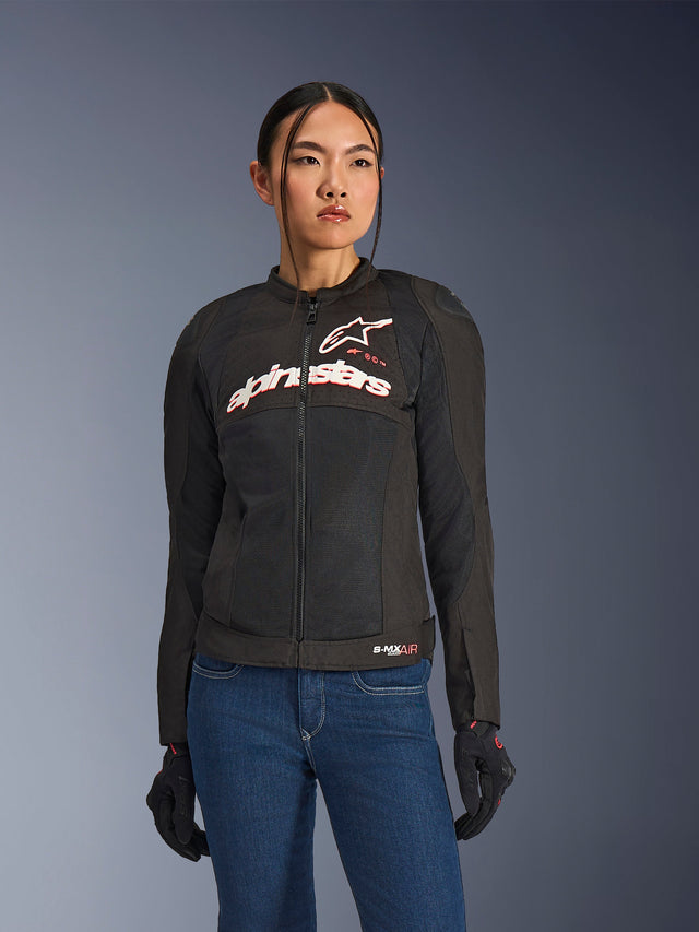 Femme portant la veste Alpinestars Stella SMX Air, noir, blanc et rouge fluo, veste de moto en textile avec maille ventilée et logo blanc sur la poitrine, associée à un jean bleu et des gants de moto noirs, coupe féminine pour la conduite sportive