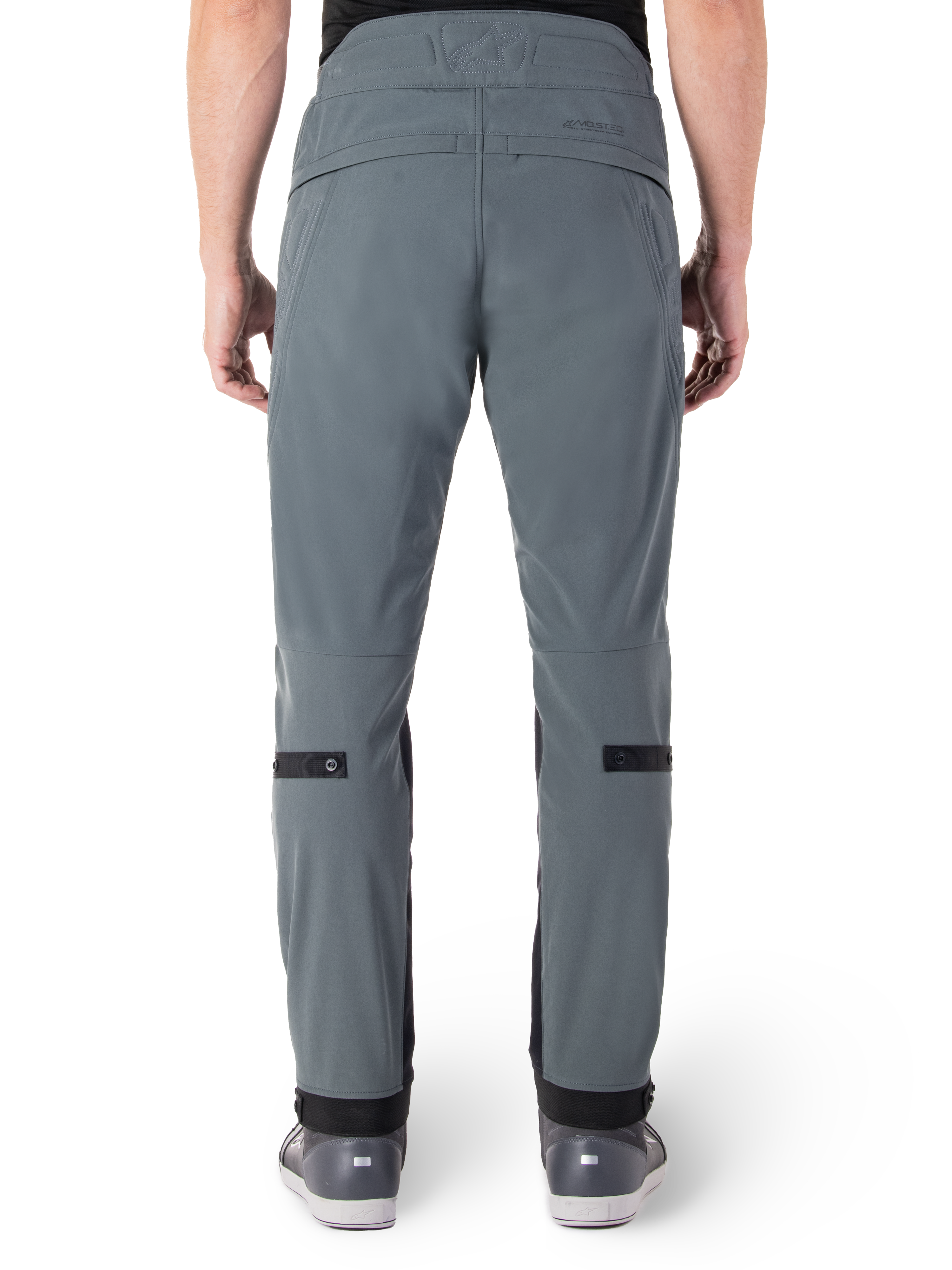 Pantalon Alpinestars Alden, Urban Gray, vue arrière d'un pantalon de moto urbain pour homme, coupe ajustée sous le genou avec des sangles de réglage noires aux mollets, design à taille haute arborant le mot-symbole MO.ST.EQ. sur l'empiècement arrière, porté avec des chaussures de moto grises