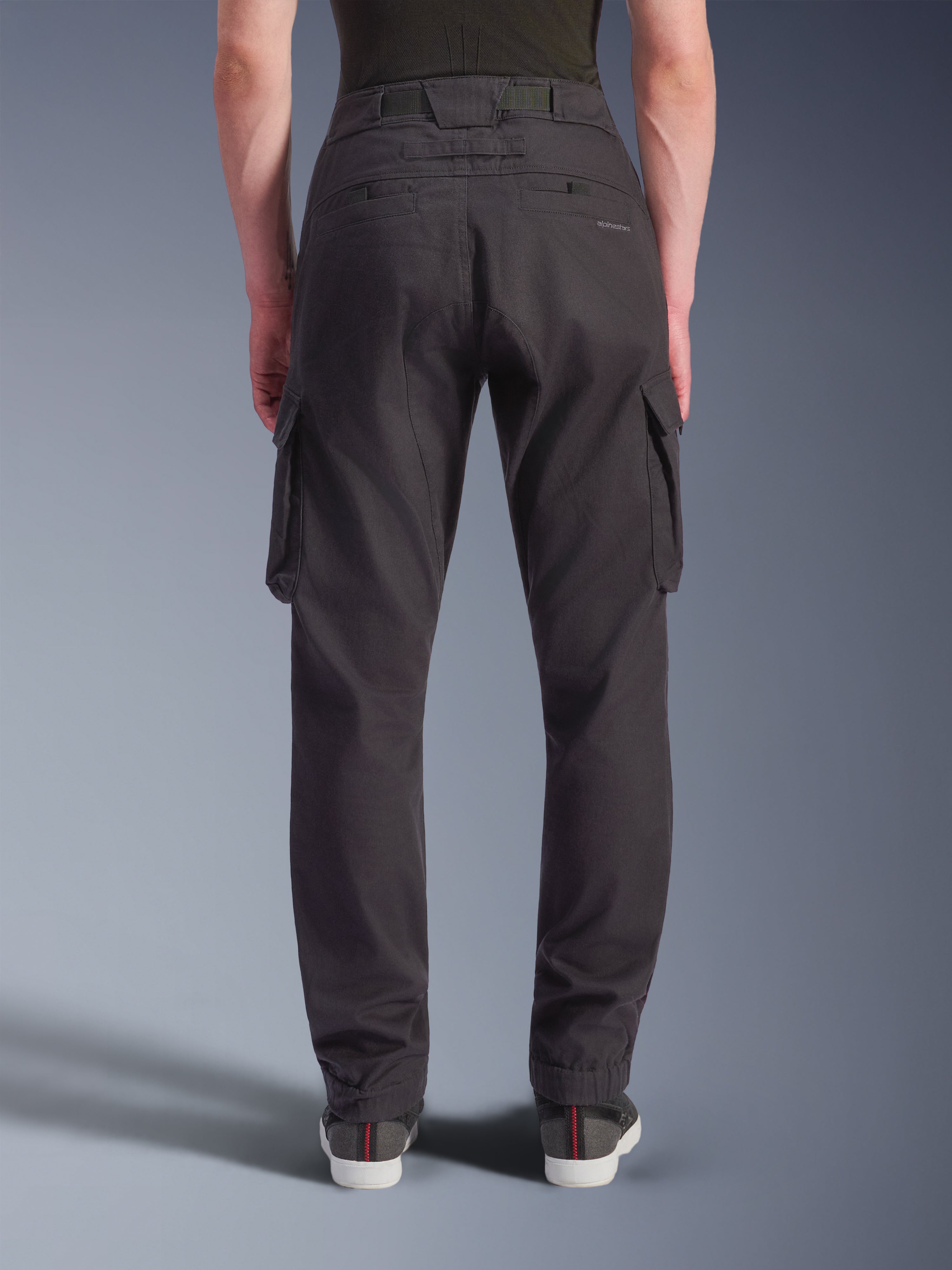 Alpinestars Flex-AST Cargo Pants - Court, pantalon de moto urbain, noir, vue arrière montrant une coupe fuselée avec ourlets élastiqués, grandes poches cargo latérales et marquage discret sur la poche arrière, porté avec des baskets décontractées