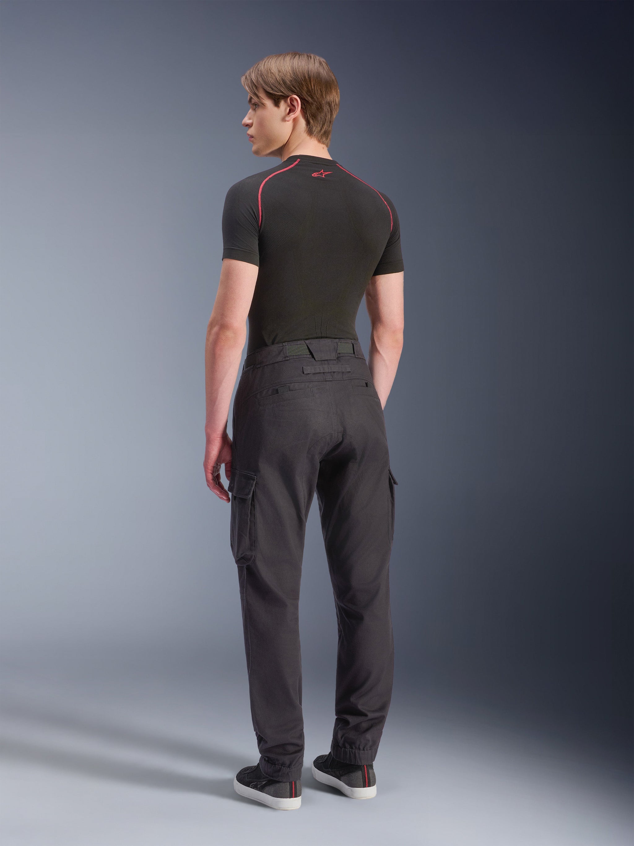 Alpinestars Flex-AST Cargo Pants, noir, pantalon de moto urbain vu de dos, avec poches cargo et jambes fuselées à ourlets élastiqués, porté avec un haut technique noir à manches courtes et des baskets noires, design technique minimaliste