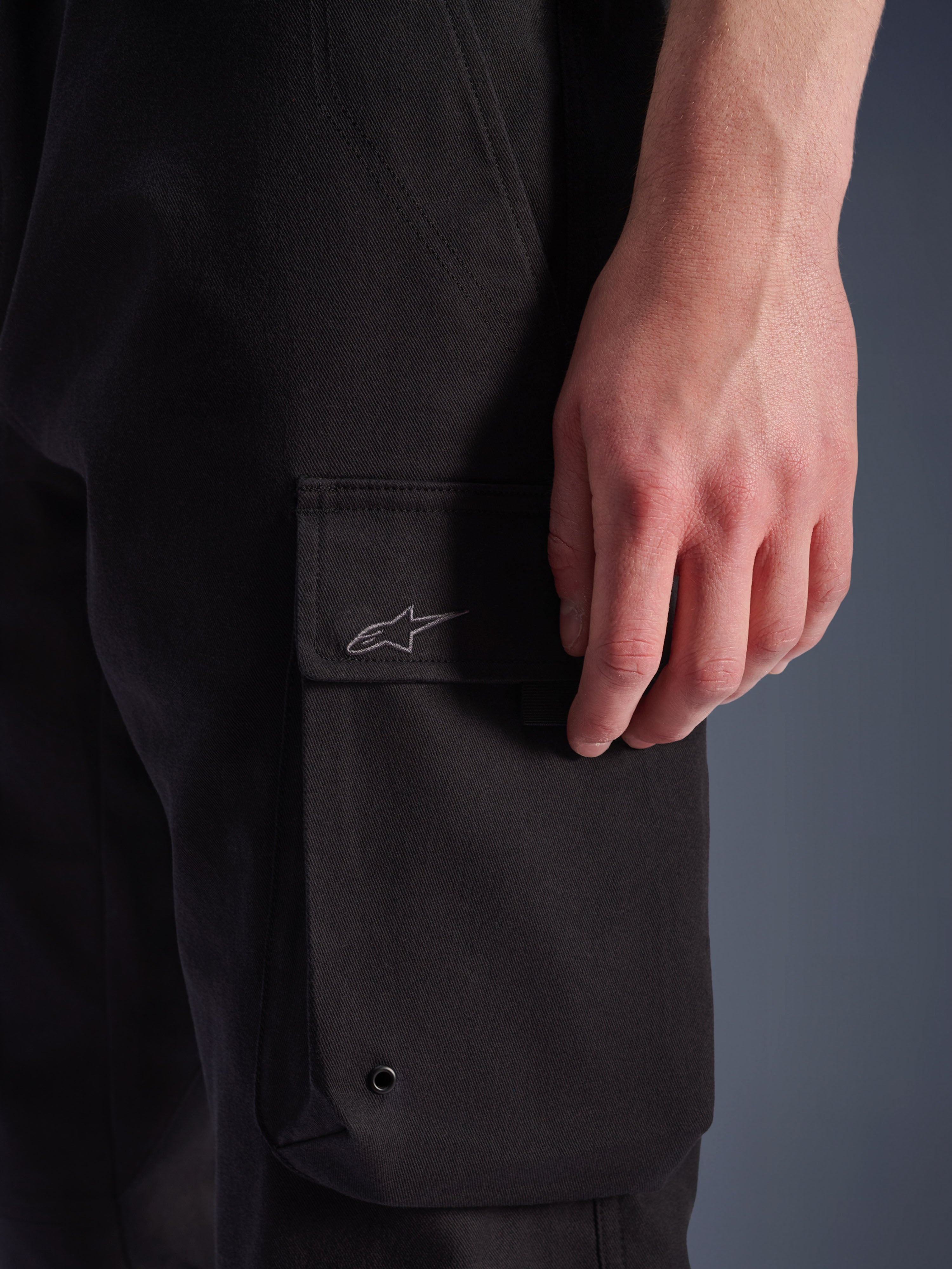 Pantalon Cargo Alpinestars Flex-AST, pantalon de moto urbain, noir, gros plan sur la poche cargo latérale avec logo Astar brodé, œillet métallique et texture en sergé extensible durable