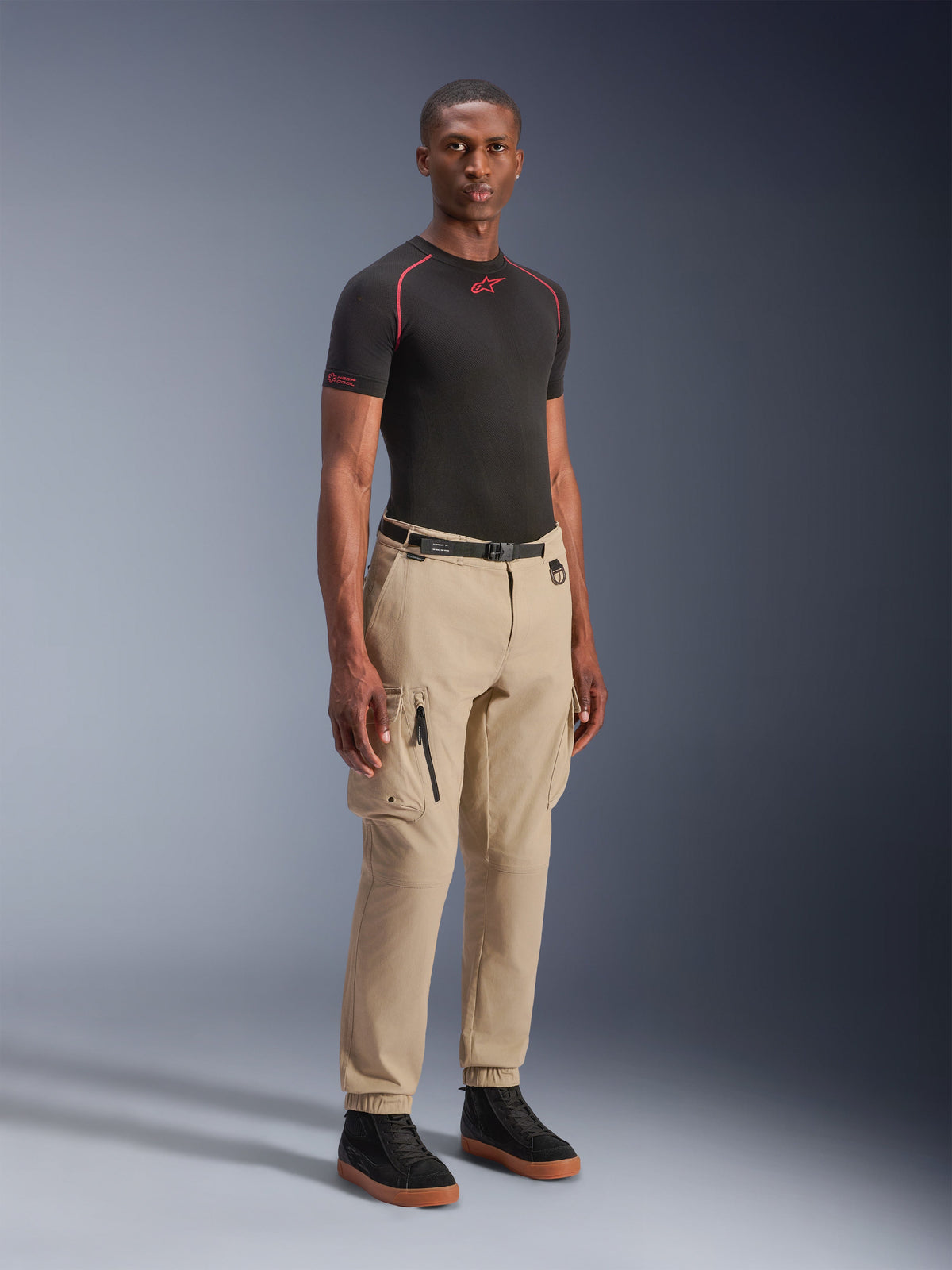 Mannequin portant le pantalon cargo Alpinestars Flex-AST en Light Beige, coupe regular tapered avec ourlets élastiqués, larges poches cargo sur les cuisses, ceinture noire, associé à une couche de base noire à manches courtes et des baskets noires, style moto urbain