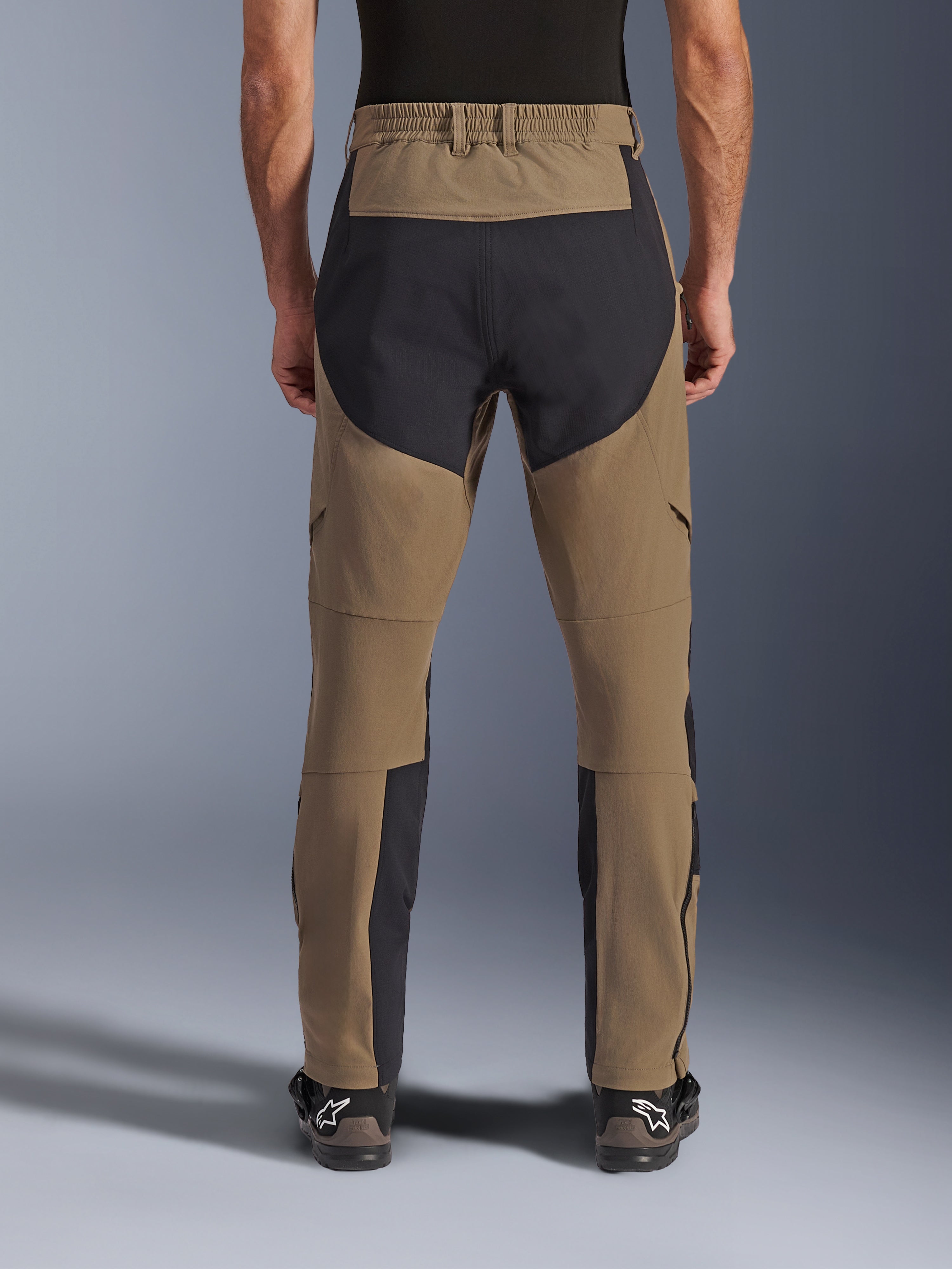 Pantalon Alpinestars Flex-AST Explorer, pantalon de moto aventure, Walnut Black, vue arrière porté par un pilote, panneaux de renfort en ripstop noir sur l'assise et l'intérieur des jambes, taille élastiquée, longues fermetures éclair au bas des jambes et bottes d'aventure assorties