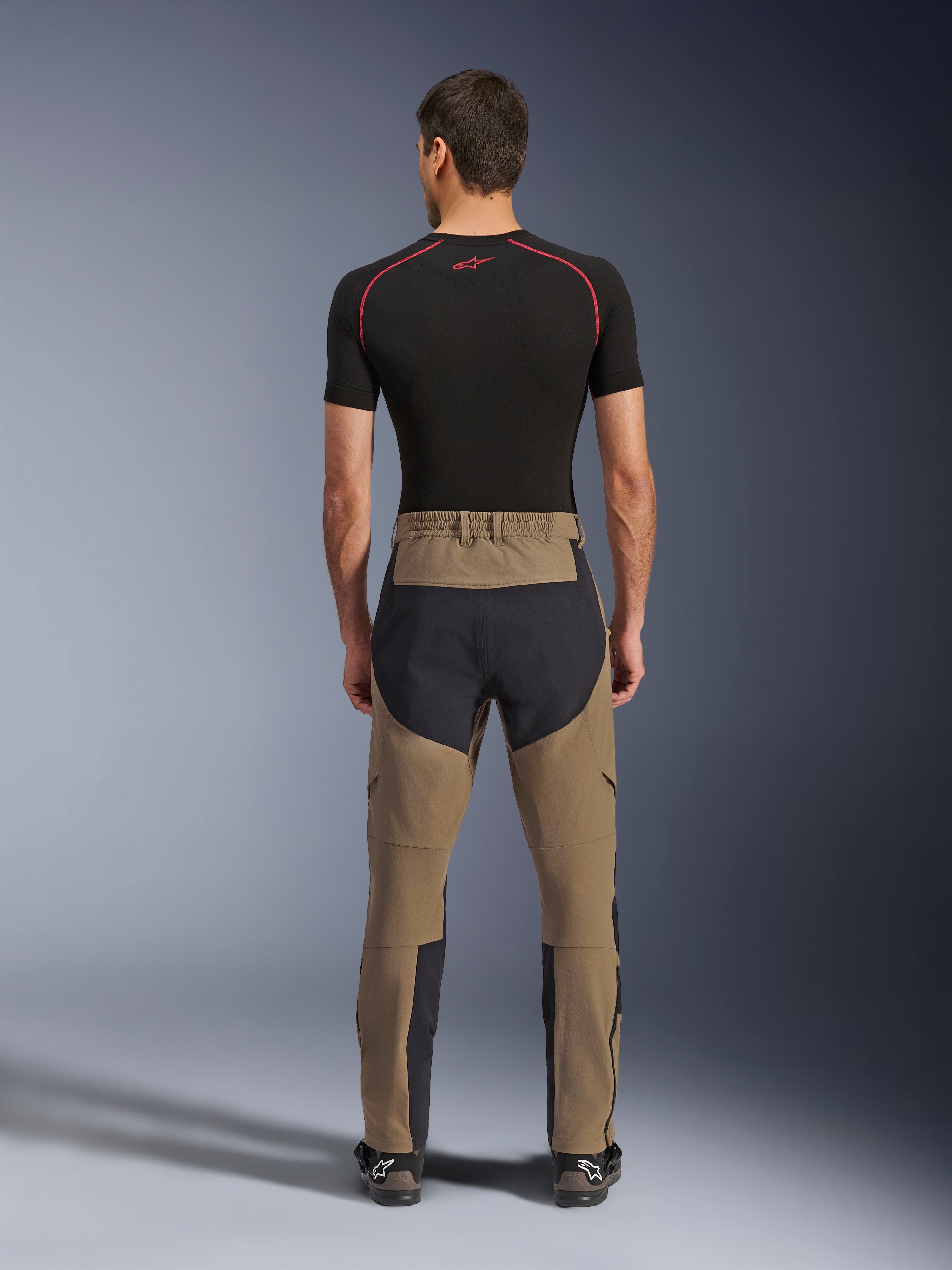 Pantalon Alpinestars Flex-AST Explorer, Walnut Black, vue arrière sur modèle, avec assise renforcée noire et textile extensible marron, associé à une couche de base noire et des bottes de moto techniques, équipement de moto d'aventure robuste