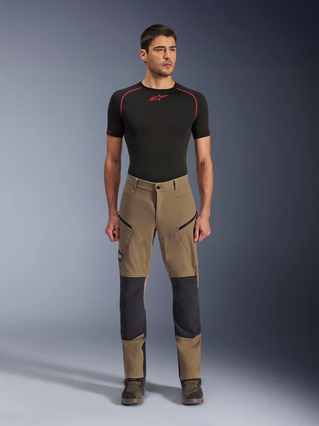 Pantalon Alpinestars Flex-AST Explorer, pantalon de moto d'aventure, Walnut Black, porté par un mannequin avec une couche de base noire et des bottes, doté d'un textile extensible beige avec des empiècements de genoux renforcés noirs et des poches cargo zippées, conçu pour le touring d'aventure.