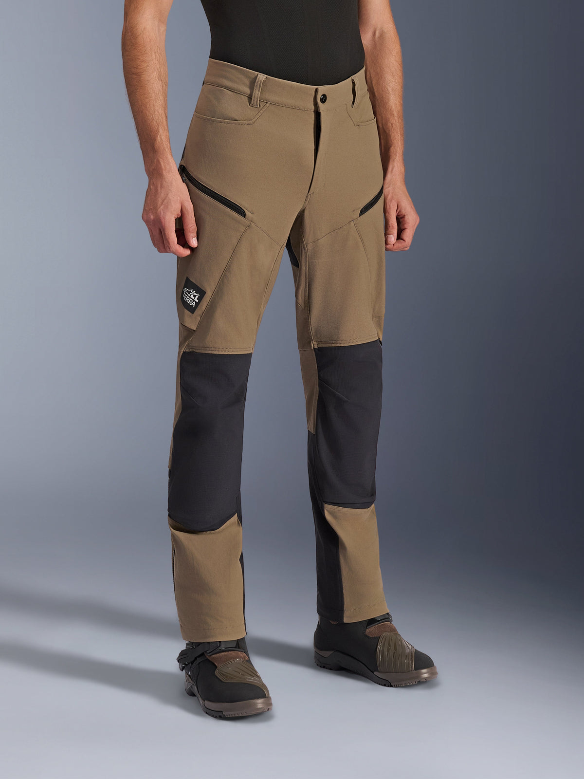 Pantalon Alpinestars Flex-AST Explorer, pantalon de moto d'aventure en Walnut Black, textile beige avec panneaux renforcés noirs aux genoux et à l'assise, poches cargo zippées et écusson du logo All Terra, porté avec des bottes de moto techniques.