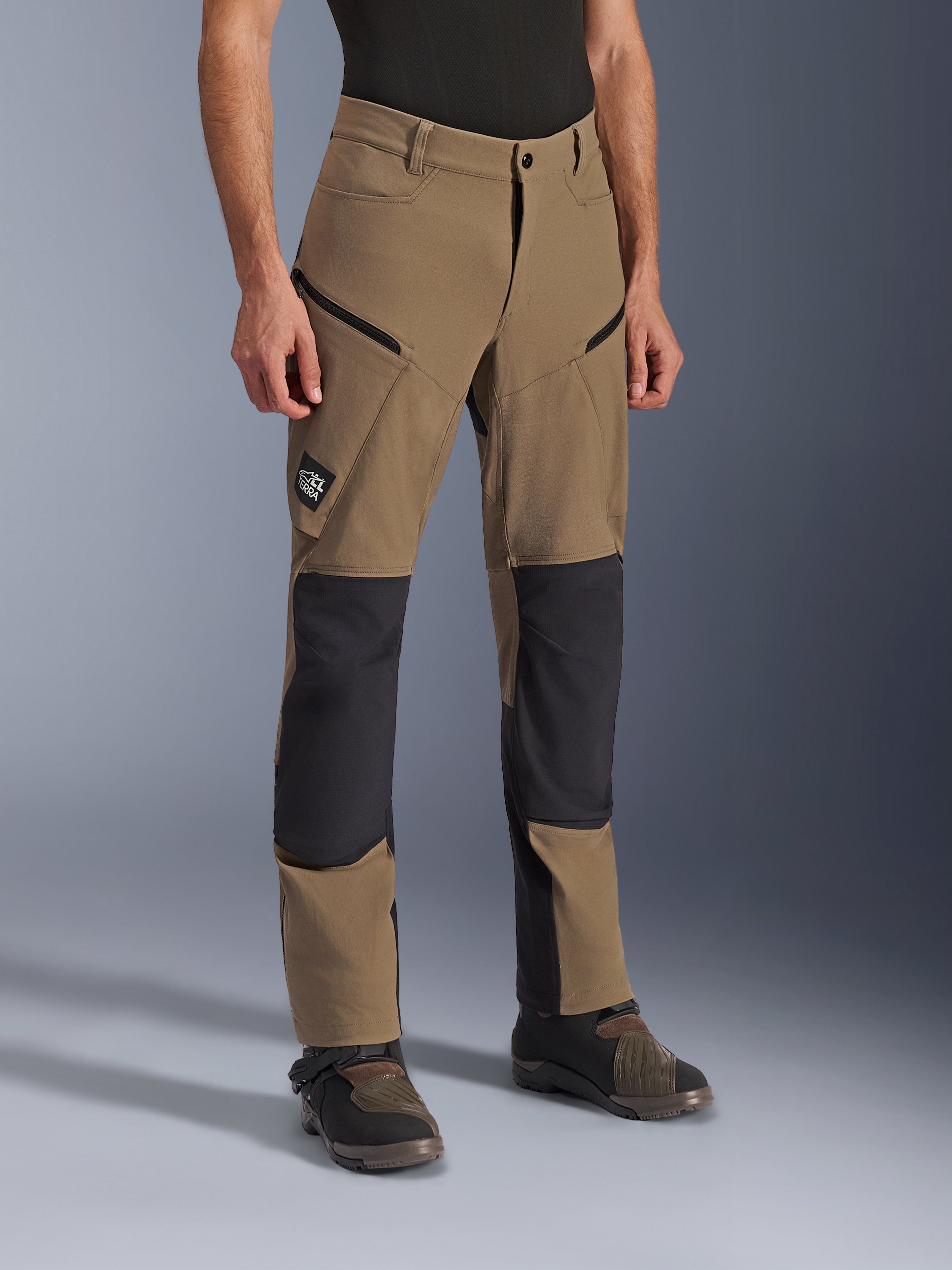 Pantalon Alpinestars Flex-AST Explorer, pantalon de moto d'aventure en Walnut Black, textile beige avec panneaux renforcés noirs aux genoux et à l'assise, poches cargo zippées et écusson du logo All Terra, porté avec des bottes de moto techniques.