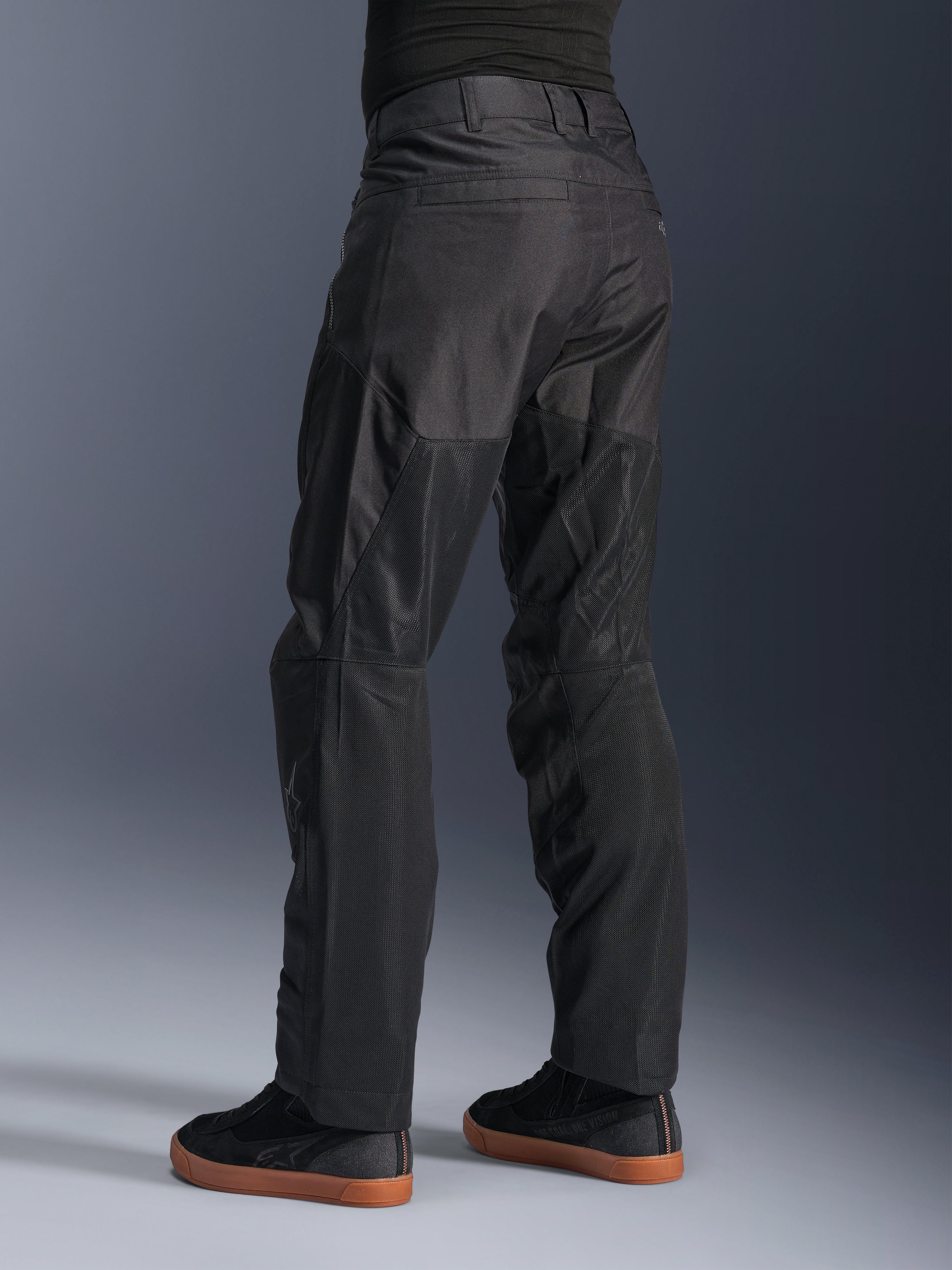 Pantalon Alpinestars C-1 Air, pantalon de moto urbain, noir, vue arrière montrant de larges panneaux en mesh technique à l'arrière des jambes pour la ventilation, style épuré pour la conduite urbaine, porté avec des baskets noires décontractées.