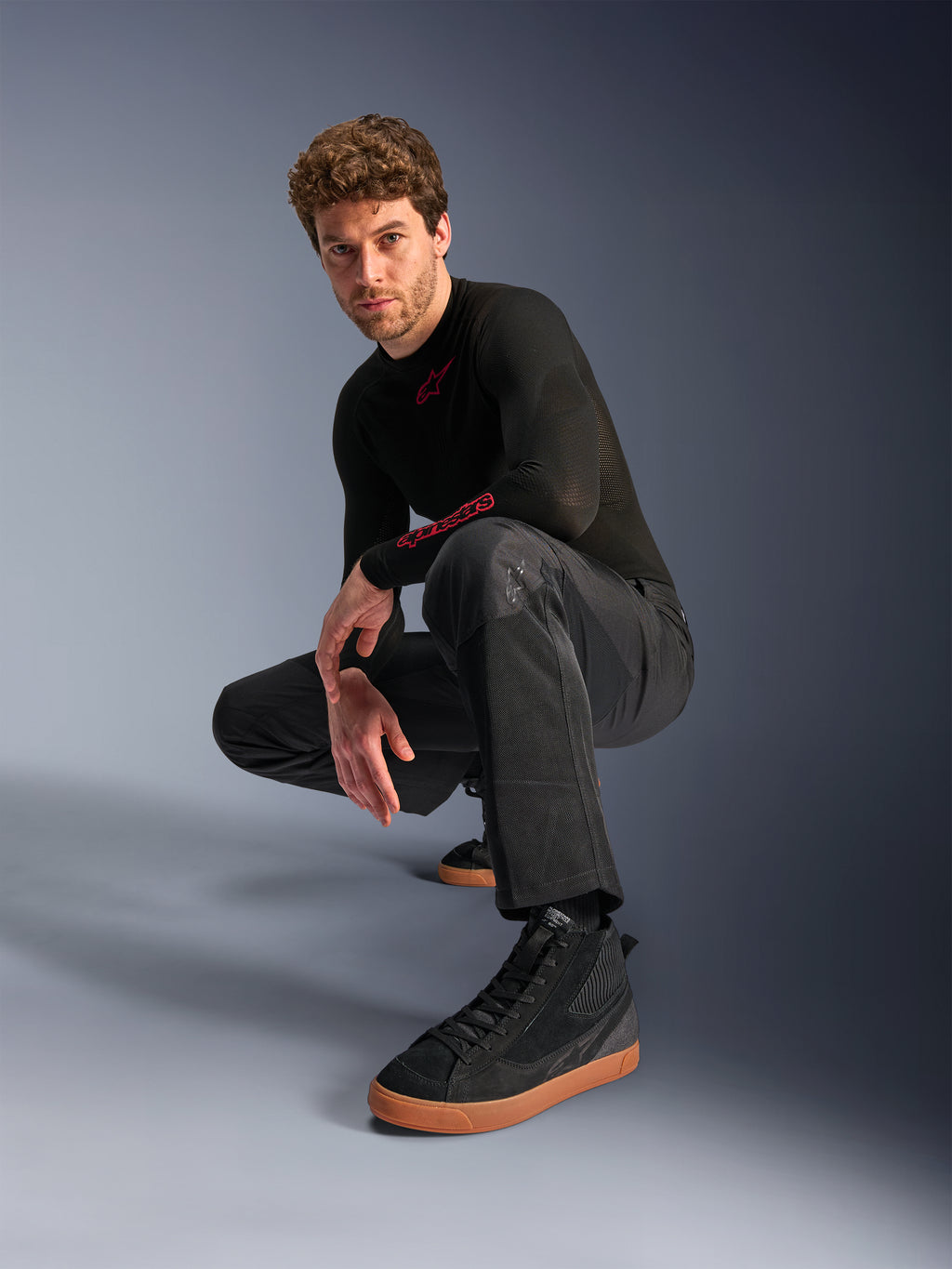 Alpinestars C-1 Air Pants, pantalon de moto urbain, noir, modèle accroupi dans un studio, porté avec un haut noir à manches longues et des baskets noires à semelles marron, doté de panneaux en mesh respirant et d'un style urbain contemporain