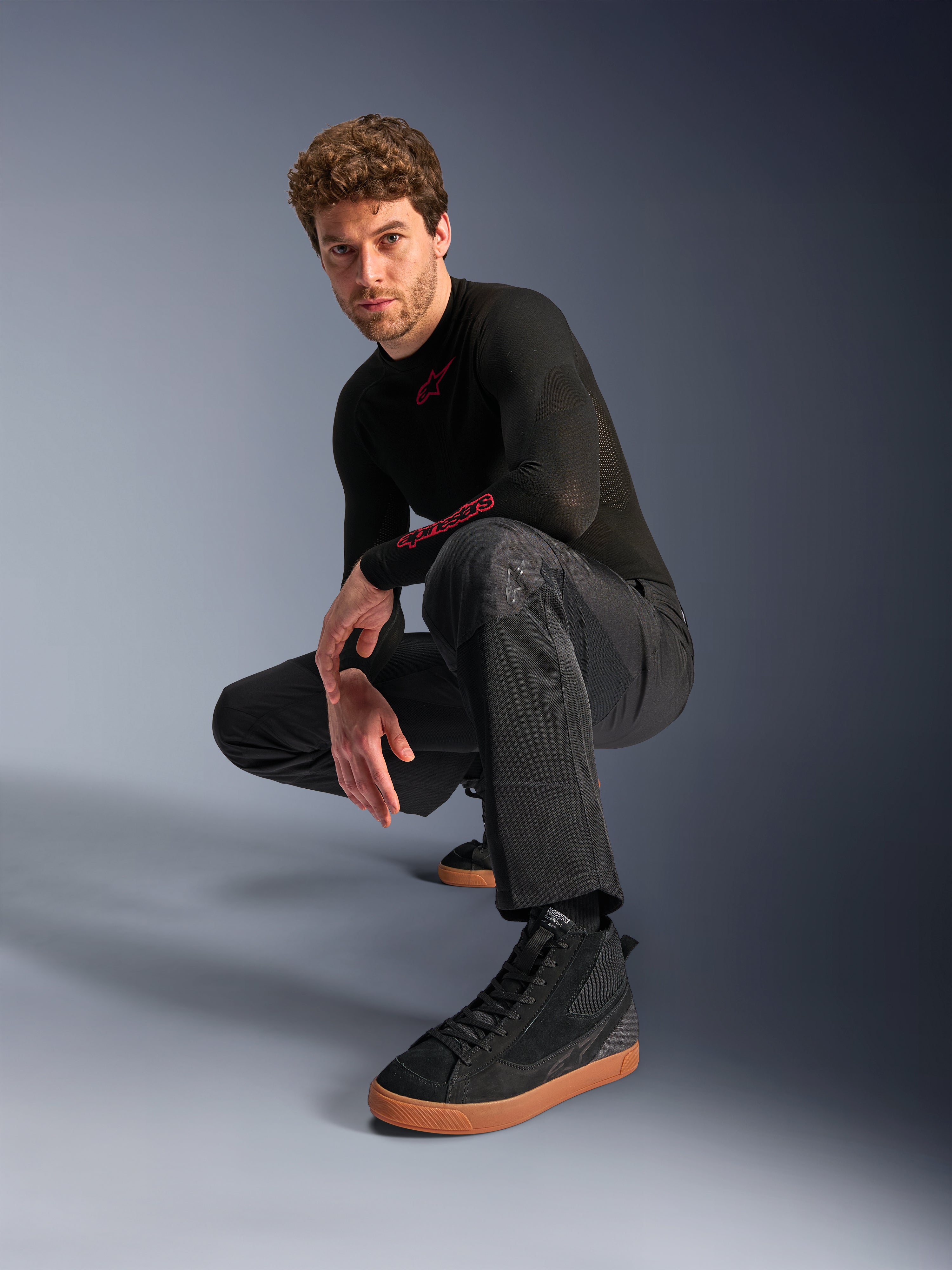Alpinestars C-1 Air Pants, pantalon de moto urbain, noir, modèle accroupi dans un studio, porté avec un haut noir à manches longues et des baskets noires à semelles marron, doté de panneaux en mesh respirant et d'un style urbain contemporain