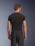 Rear view of a model wearing Alpinestars Aeroshell Airflow Pants, Black, lightweight softshell construction with integrated mesh side panels, paired with a black base layer featuring red contrast stitching, sporty motorcycle attire