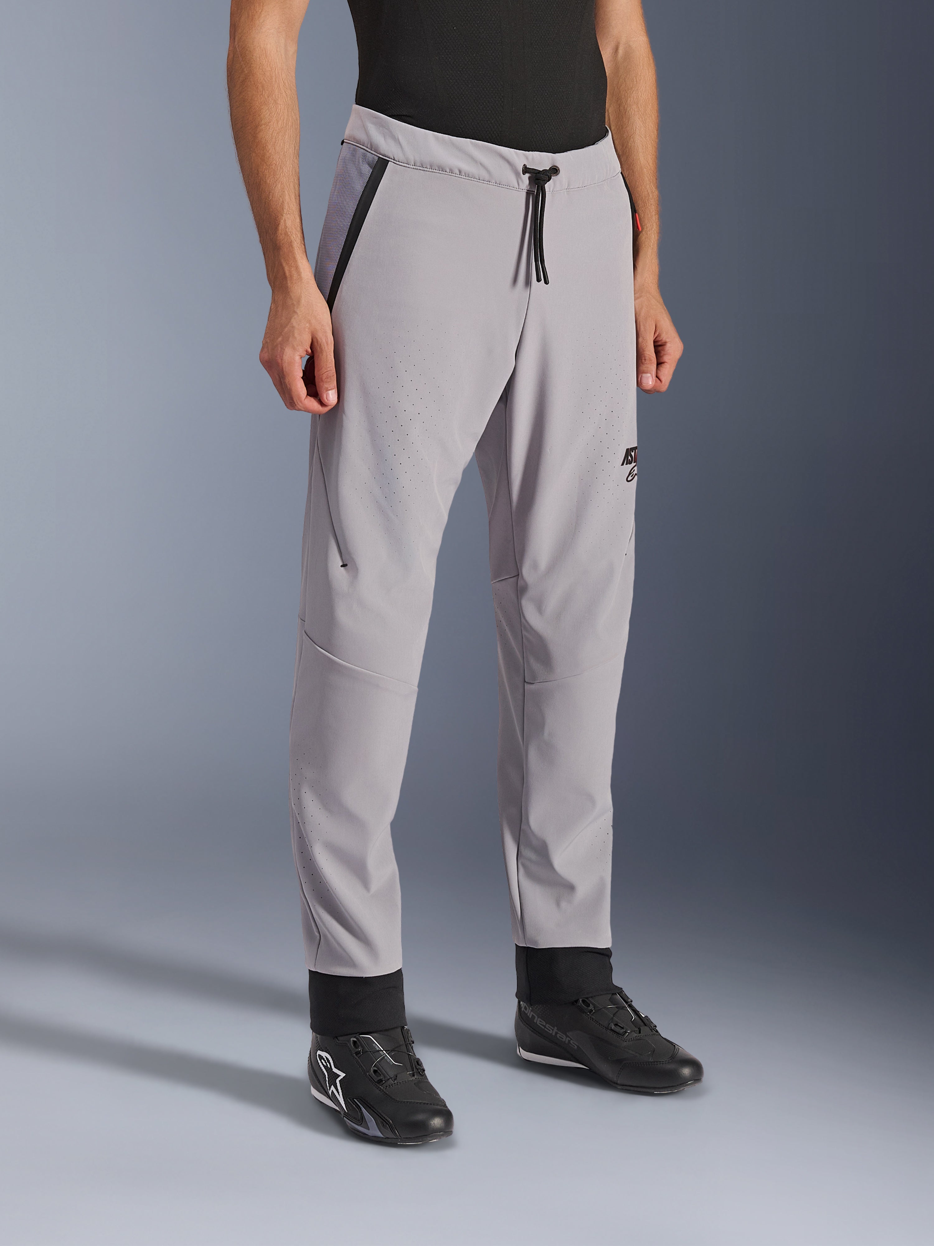 Pantalon Alpinestars Aeroshell Airflow, gris moyen, pantalon de moto léger avec zones de ventilation perforées au laser sur les cuisses et les tibias, doté d'une taille élastique avec cordon de serrage et d'une coupe sport ajustée, porté par un mannequin avec des chaussures de moto noires.