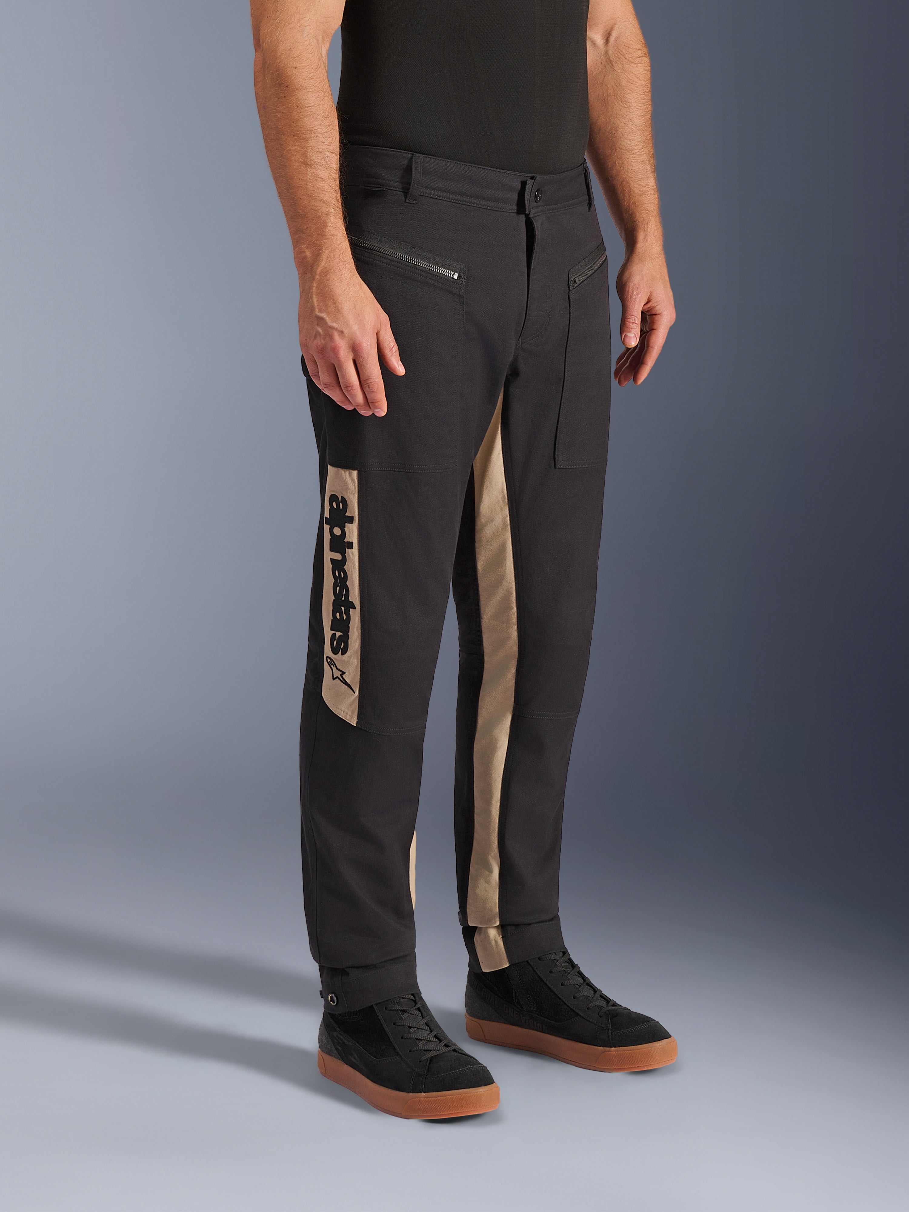 Pantalon Alpinestars Flex-AST Canvas, pantalon de moto urbain, Black Aluminum, doté de panneaux latéraux beiges avec un logo Alpinestars vertical, design inspiré du workwear avec poches zippées et coupe ergonomique, porté avec des baskets noires décontractées pour un look urbain moderne.
